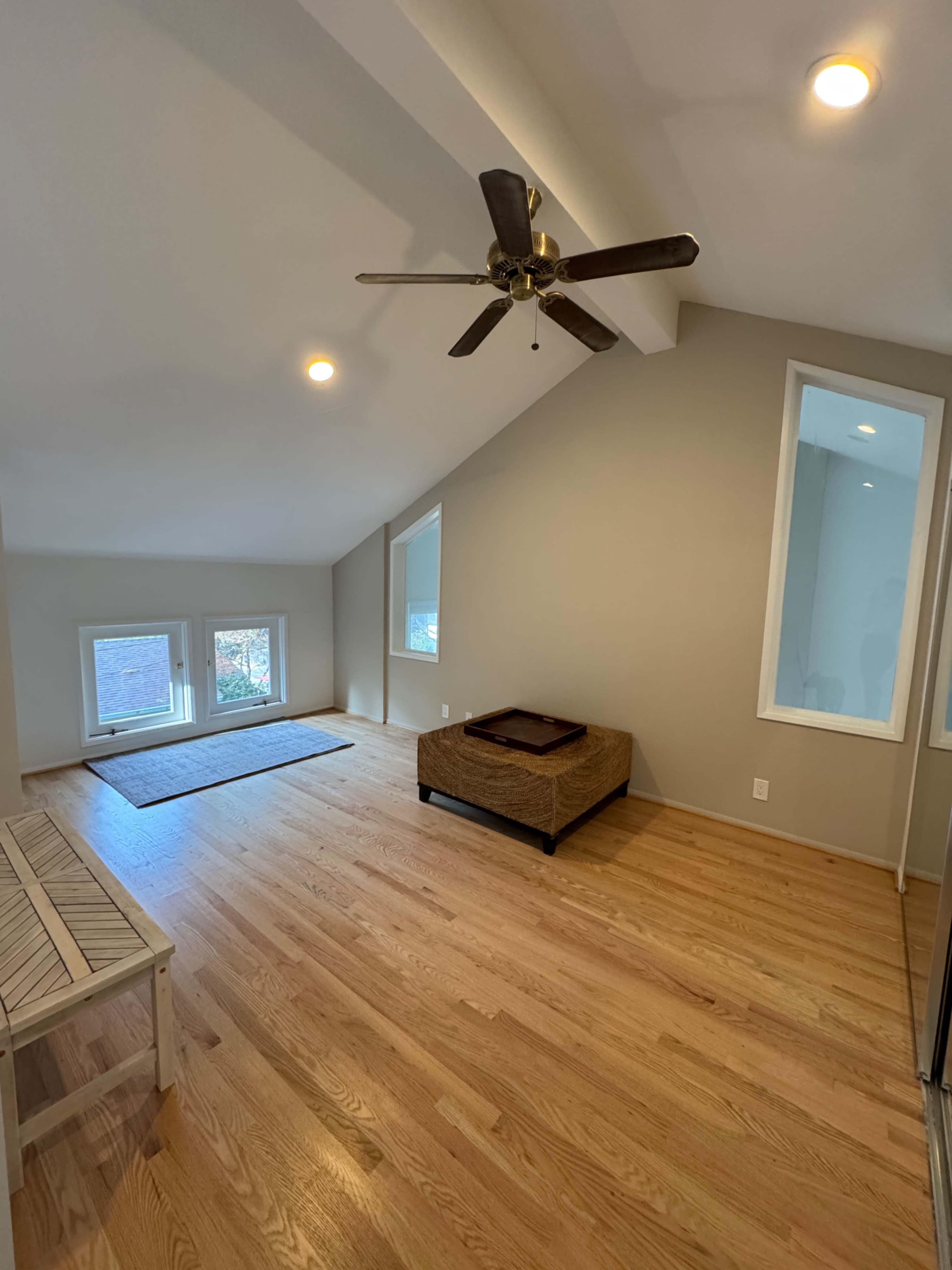 The image shows a spacious, light-filled room with hardwood floors, a ceiling fan, and two large windows facing the outdoors.