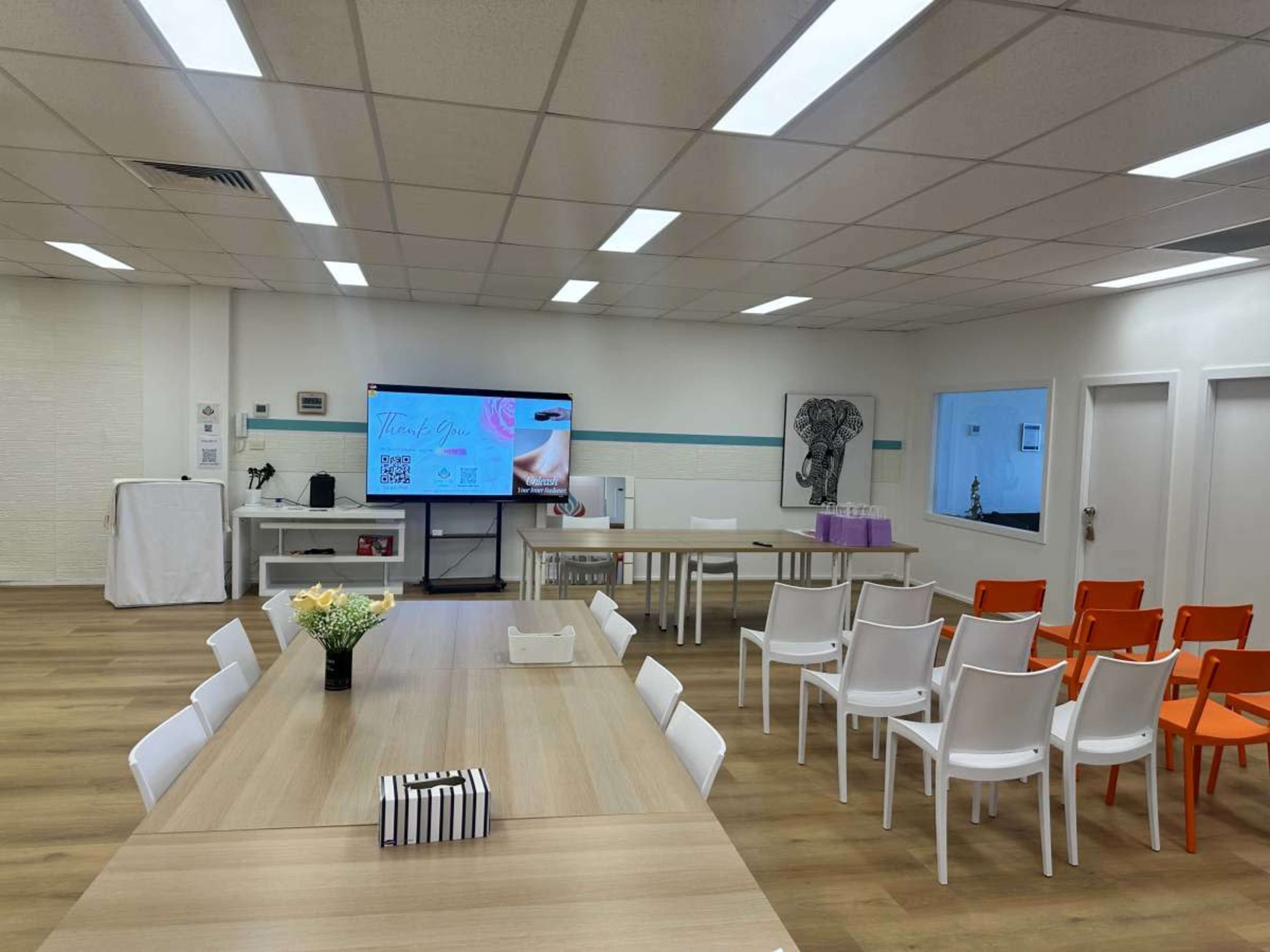 The image shows a spacious meeting room with a large screen displaying information, a long table surrounded by white chairs, and additional orange chairs along the walls.