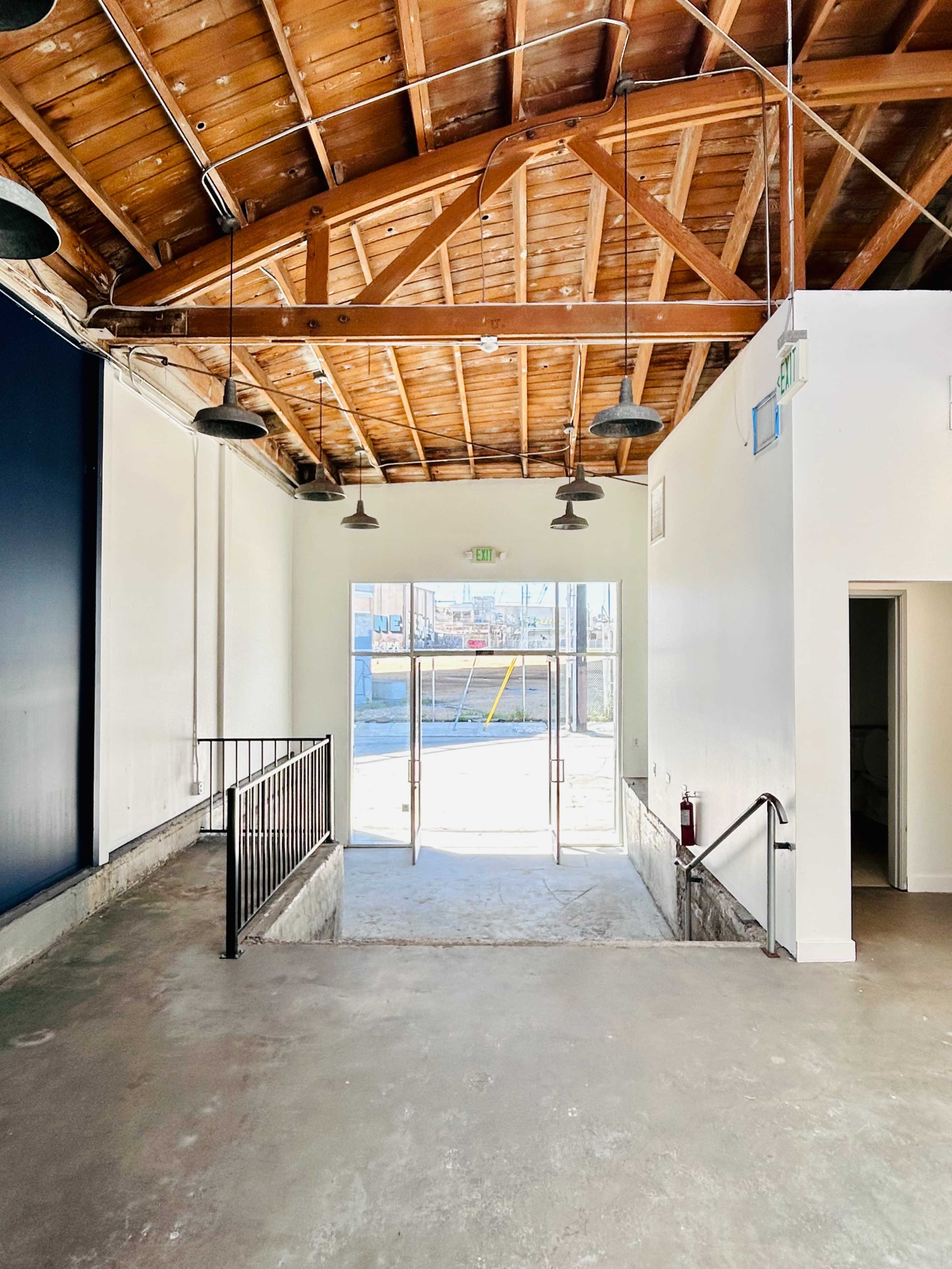 The image shows an empty commercial interior space with a wooden beam ceiling, an open entrance, and concrete flooring.