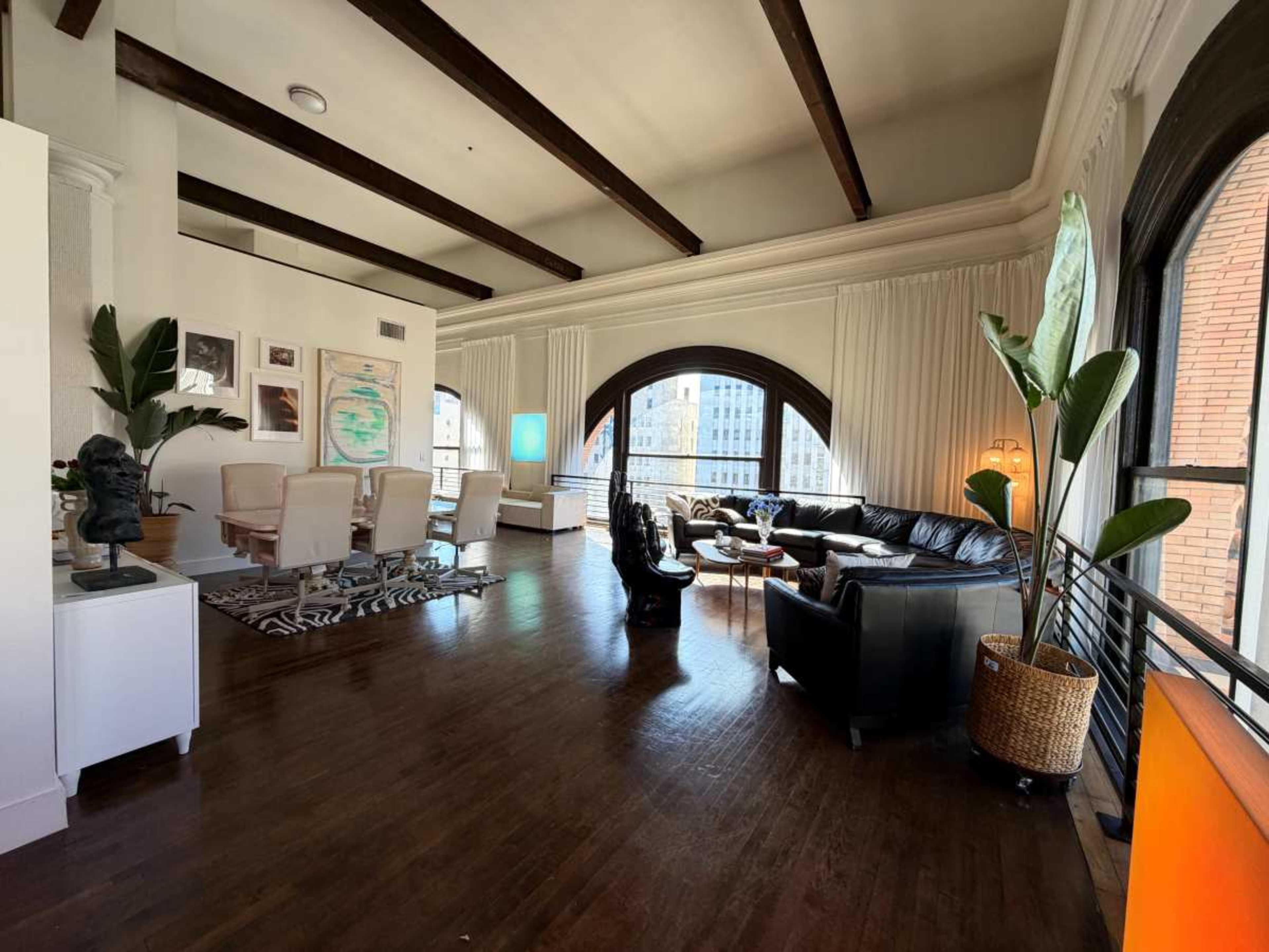 The image shows a spacious living area with large windows, featuring a mix of modern furniture and decorative plants.