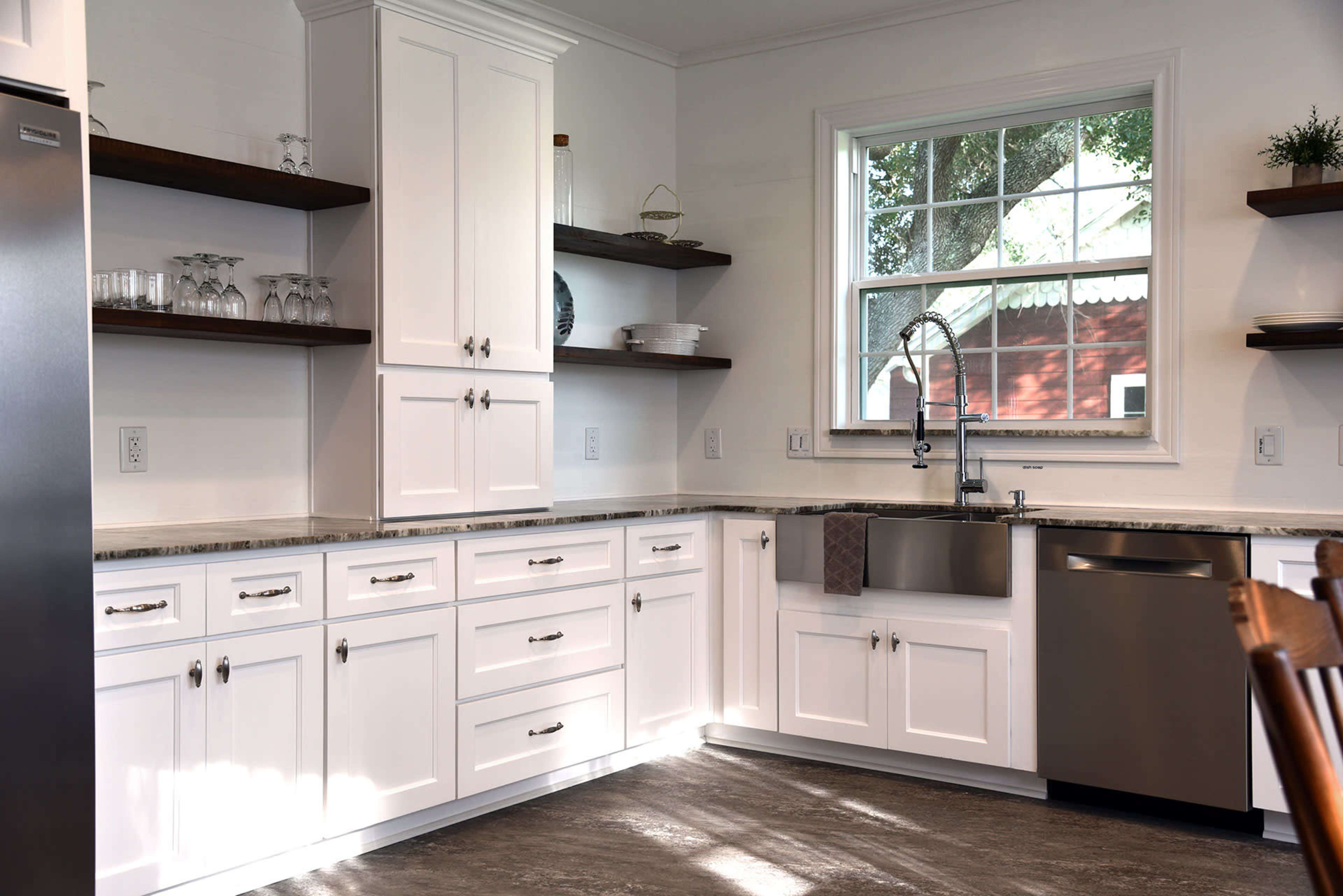 A modern kitchen with white cabinetry, open wooden shelves, and stainless steel appliances, featuring a large window that lets in natural light.