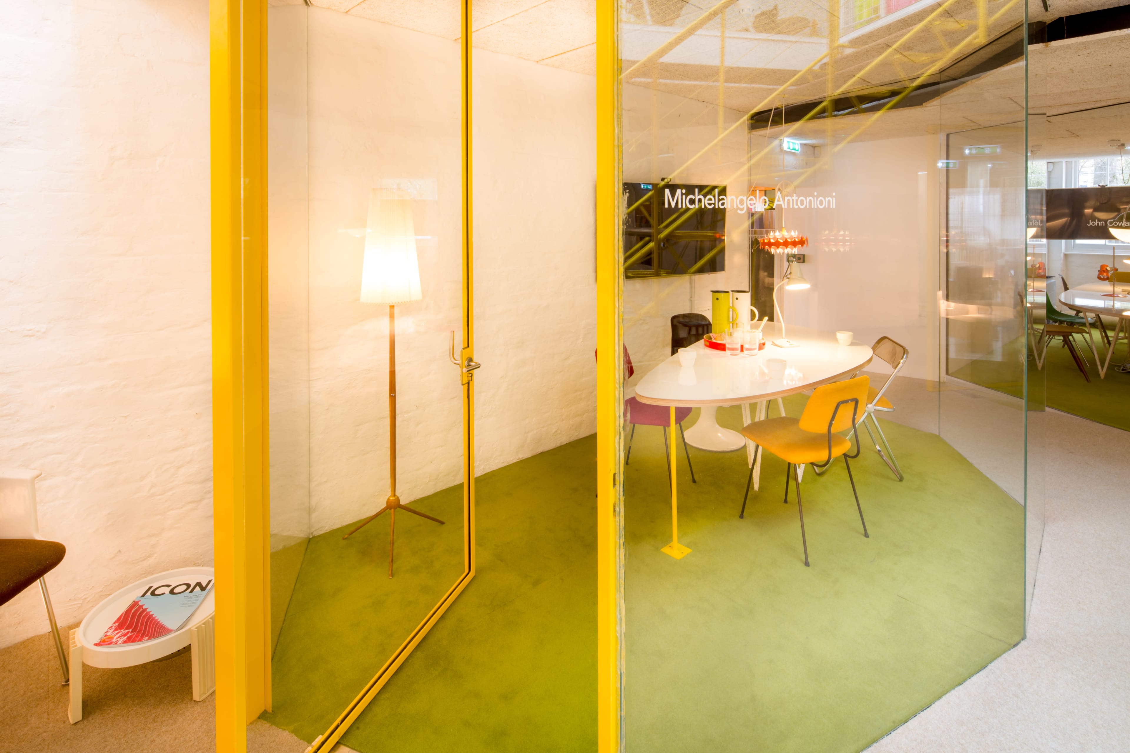 A glass-walled office space features a round white table surrounded by colorful chairs on a green carpet.