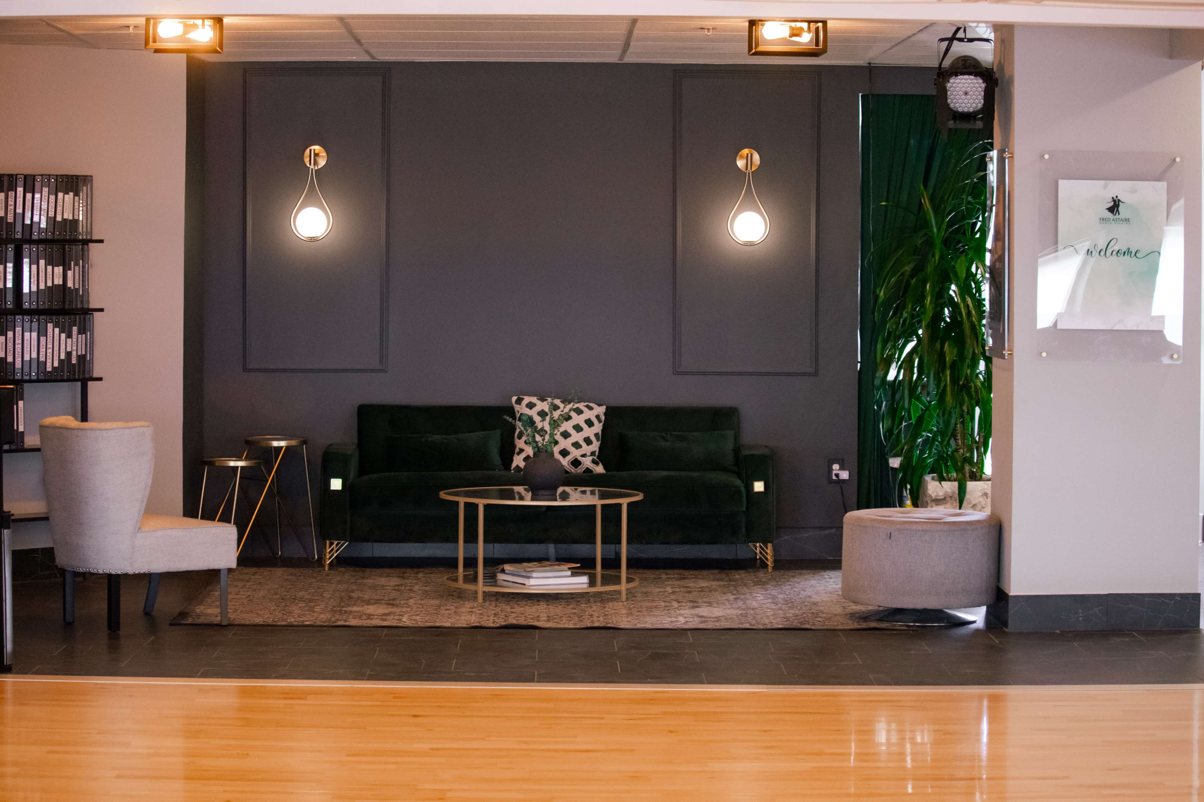 The image shows a modern lounge area with a dark green couch, a gold coffee table, and stylish lighting fixtures against a gray wall, accompanied by a decorative plant.