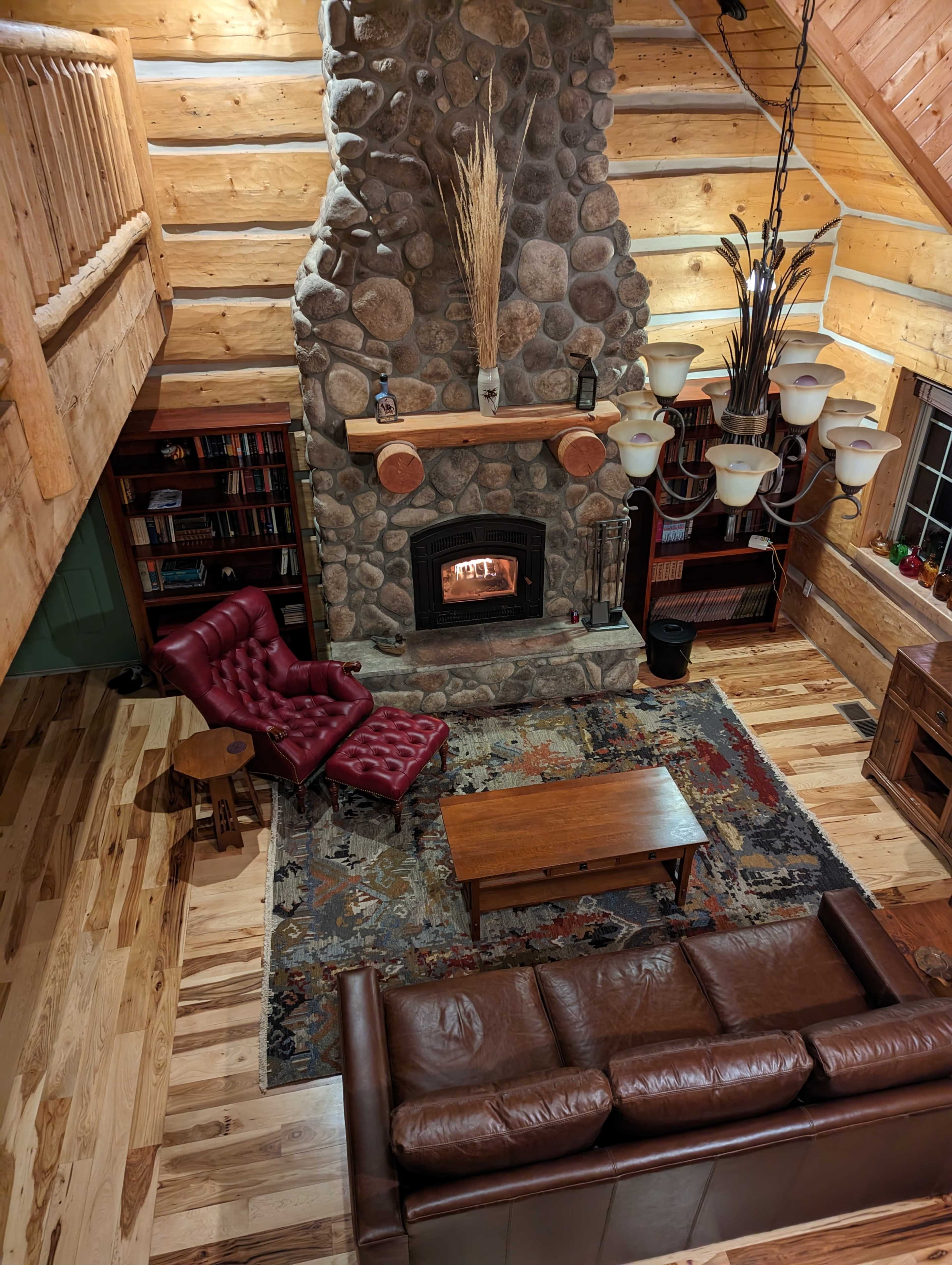 A cozy log cabin living room with a stone fireplace, a wooden coffee table, a leather sofa, and a red armchair on a patterned rug.