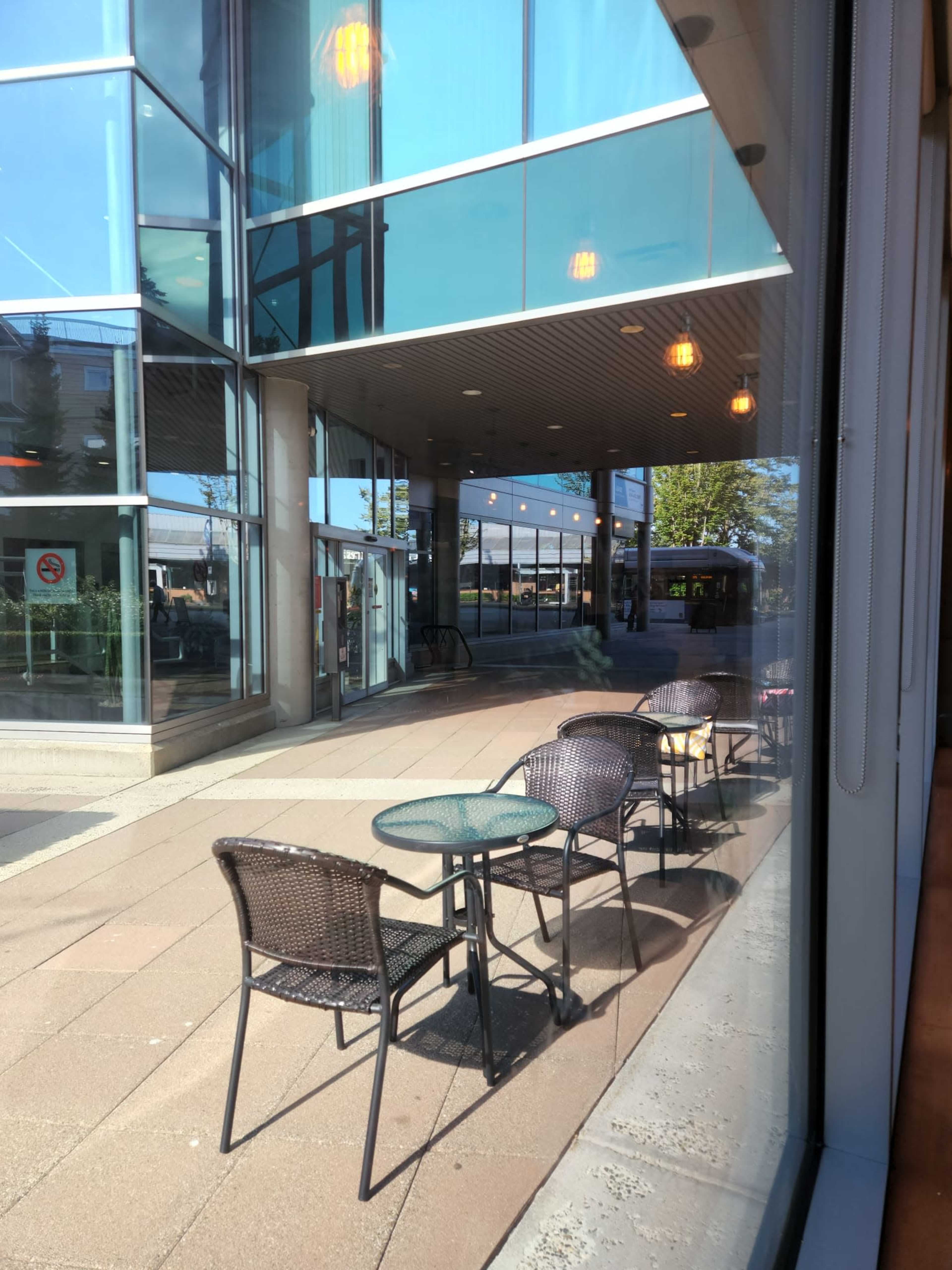 An outdoor seating area with empty tables and chairs outside a modern building, reflecting bright sunlight.