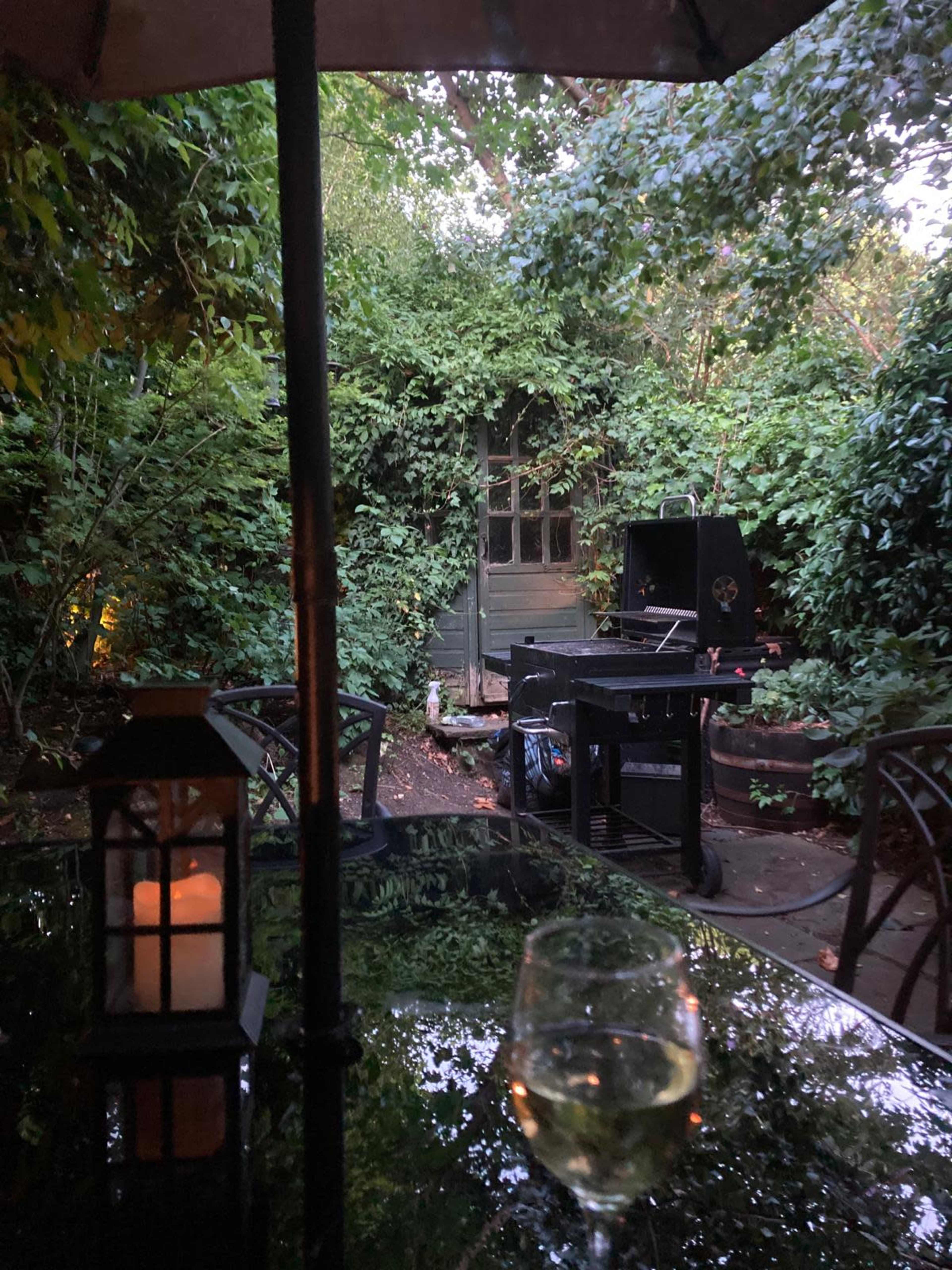 A glass of white wine rests on a table under an umbrella, with a view of a garden path obscured by dense greenery and a grill in the background.