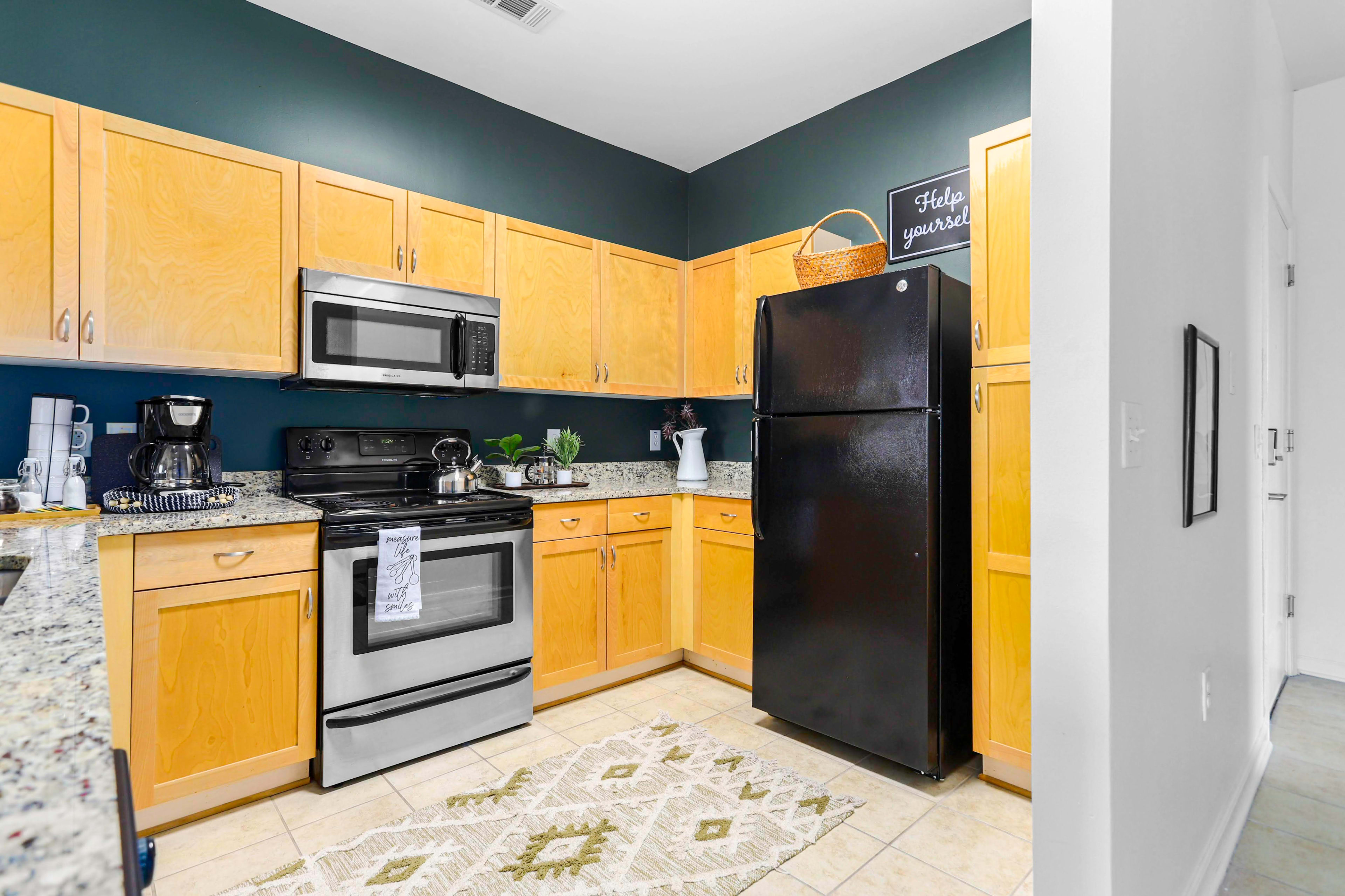 The image shows a compact kitchen with wooden cabinets, a black refrigerator, stainless steel appliances, and a granite countertop.