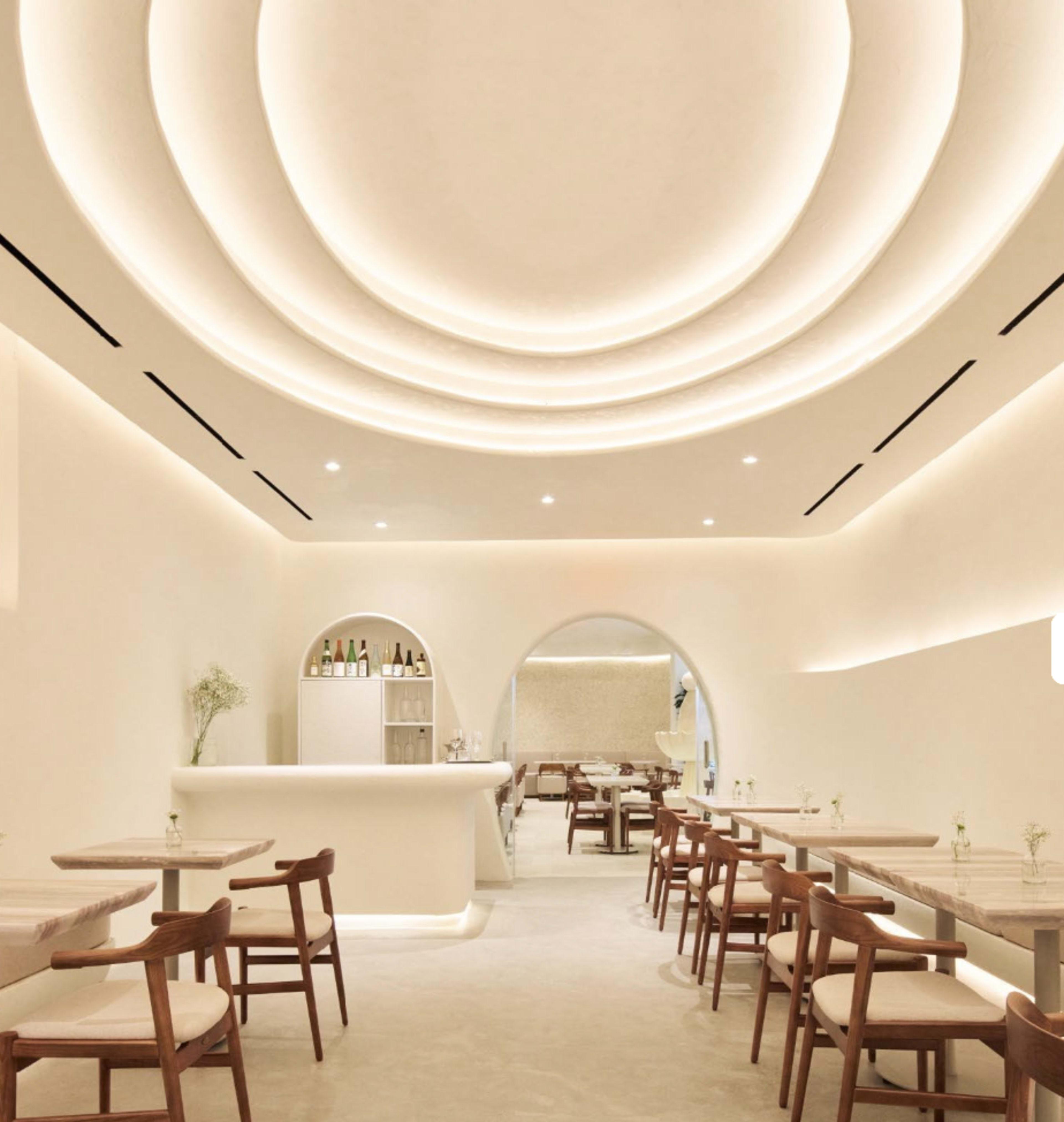 The image shows a modern, minimalist restaurant interior with arched features, soft lighting, and wooden tables and chairs arranged neatly.