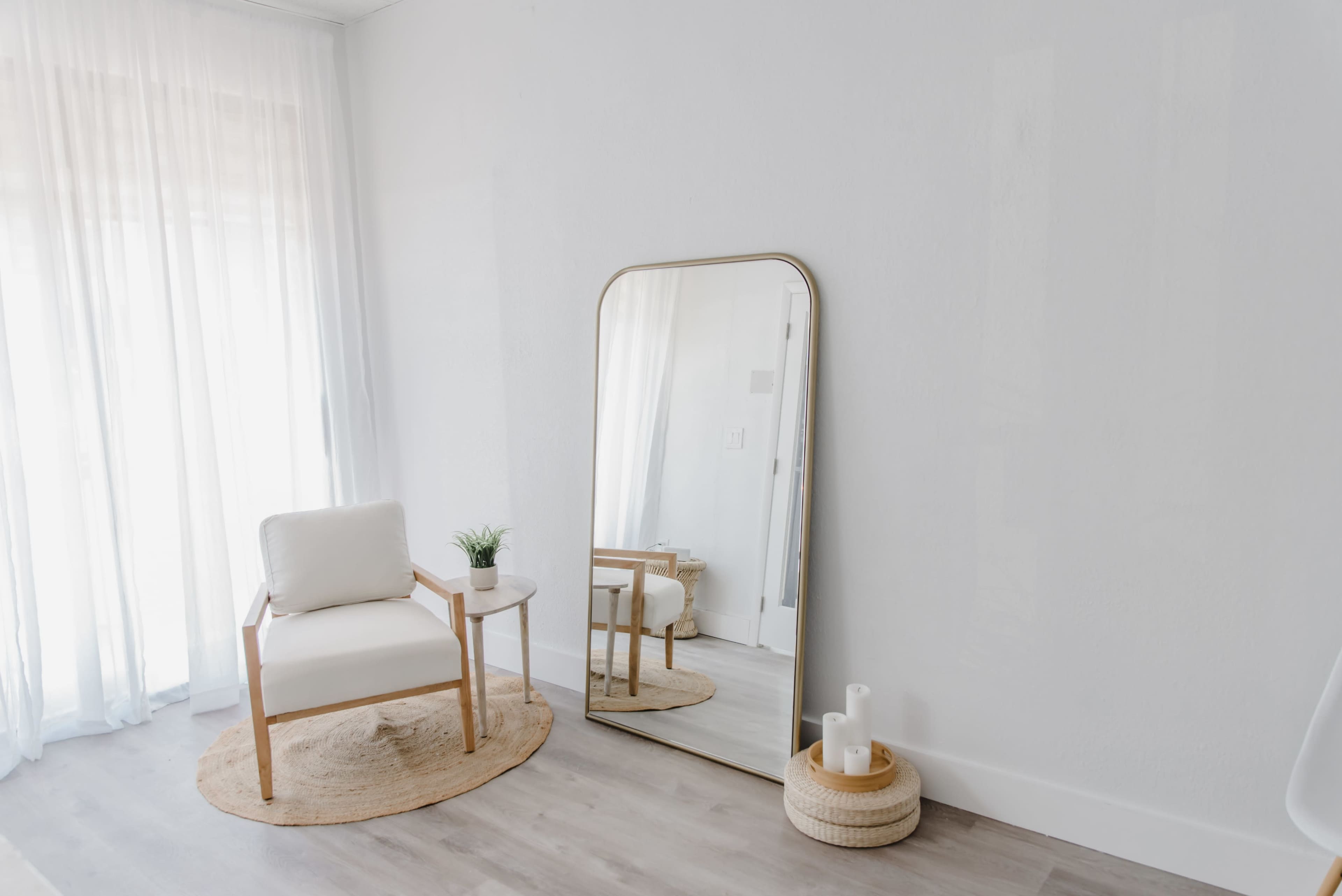 A minimalist interior features a white chair beside a round rug, a tall mirror reflecting light, and a small table with a potted plant.