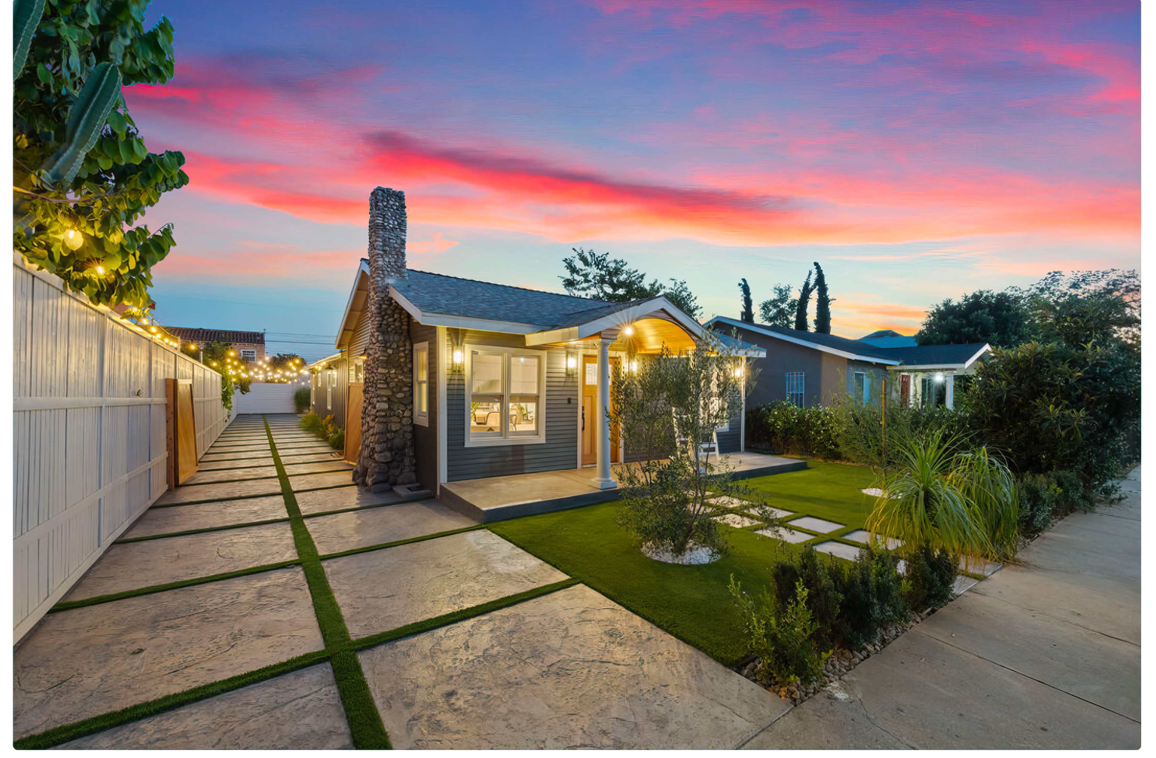 A small house with a stone chimney and landscaped front yard sits under a colorful sunset sky, flanked by walkways and greenery.