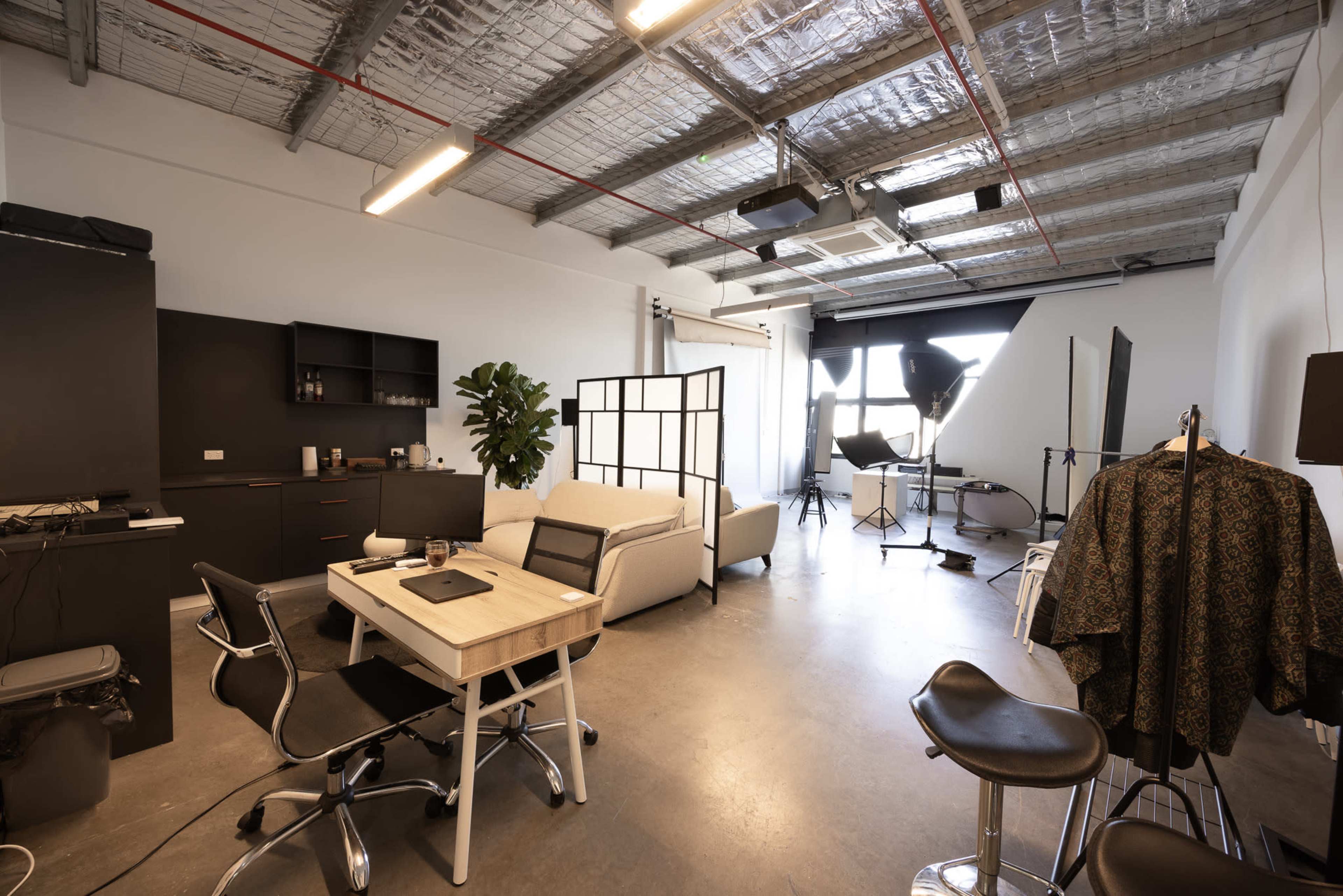 The image shows a spacious, modern studio with a variety of furniture and equipment, including a desk, chairs, a sofa, and photography lighting setups.