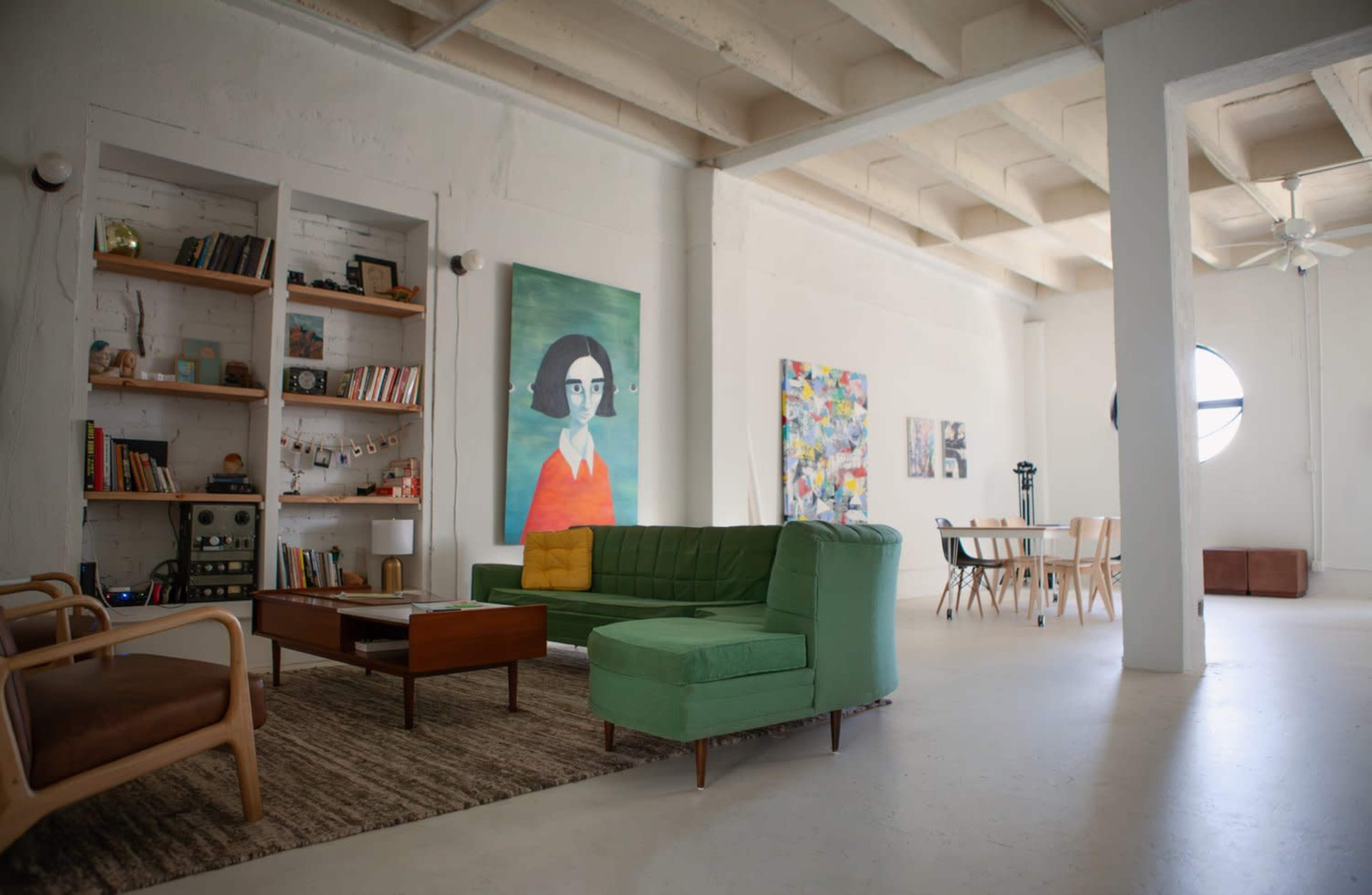 The image shows a spacious living area with a green sectional couch, wooden coffee table, bookshelves, and various artworks on the walls.