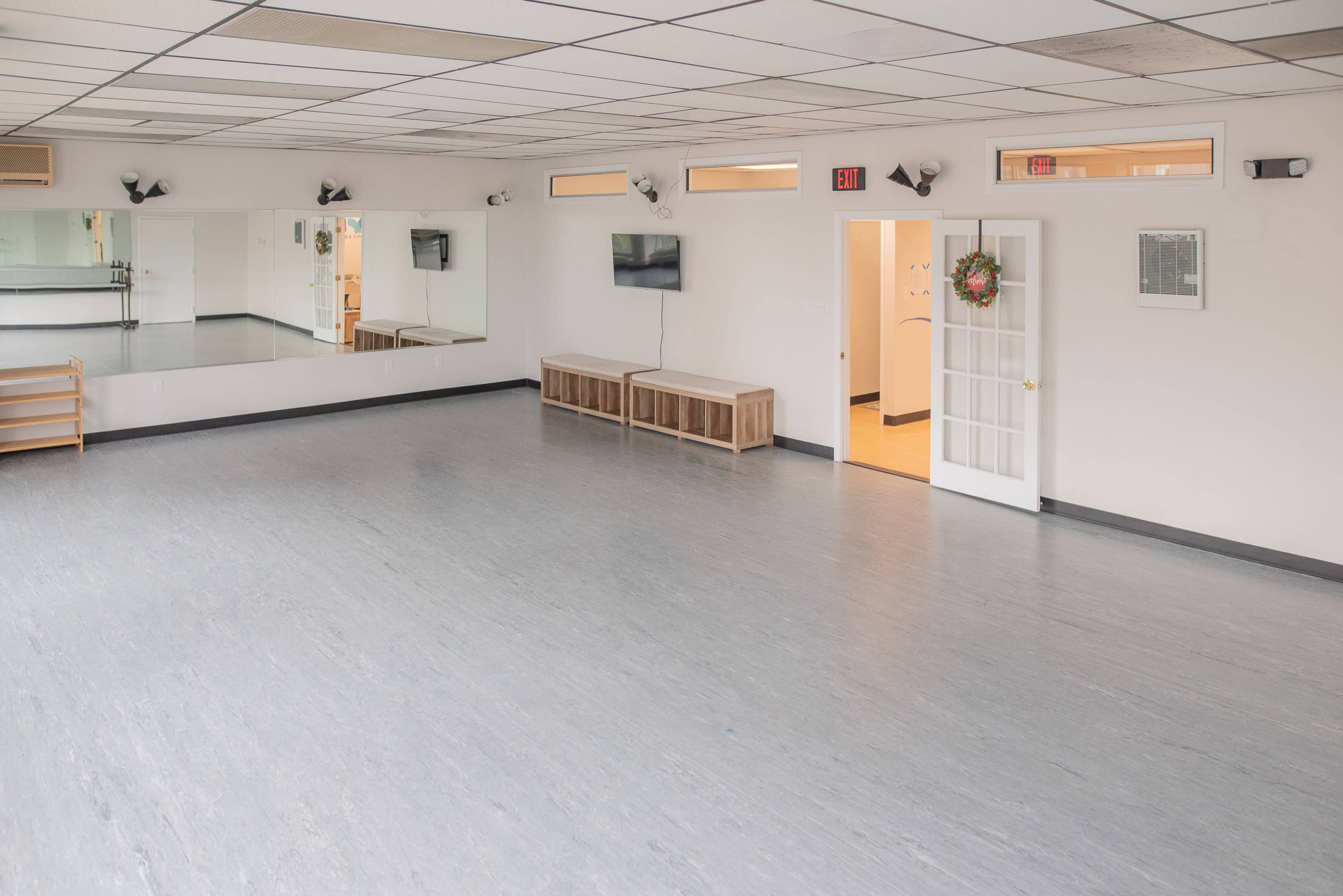 A spacious, empty dance studio with large mirrors, wooden benches, and a door leading to a restroom area.