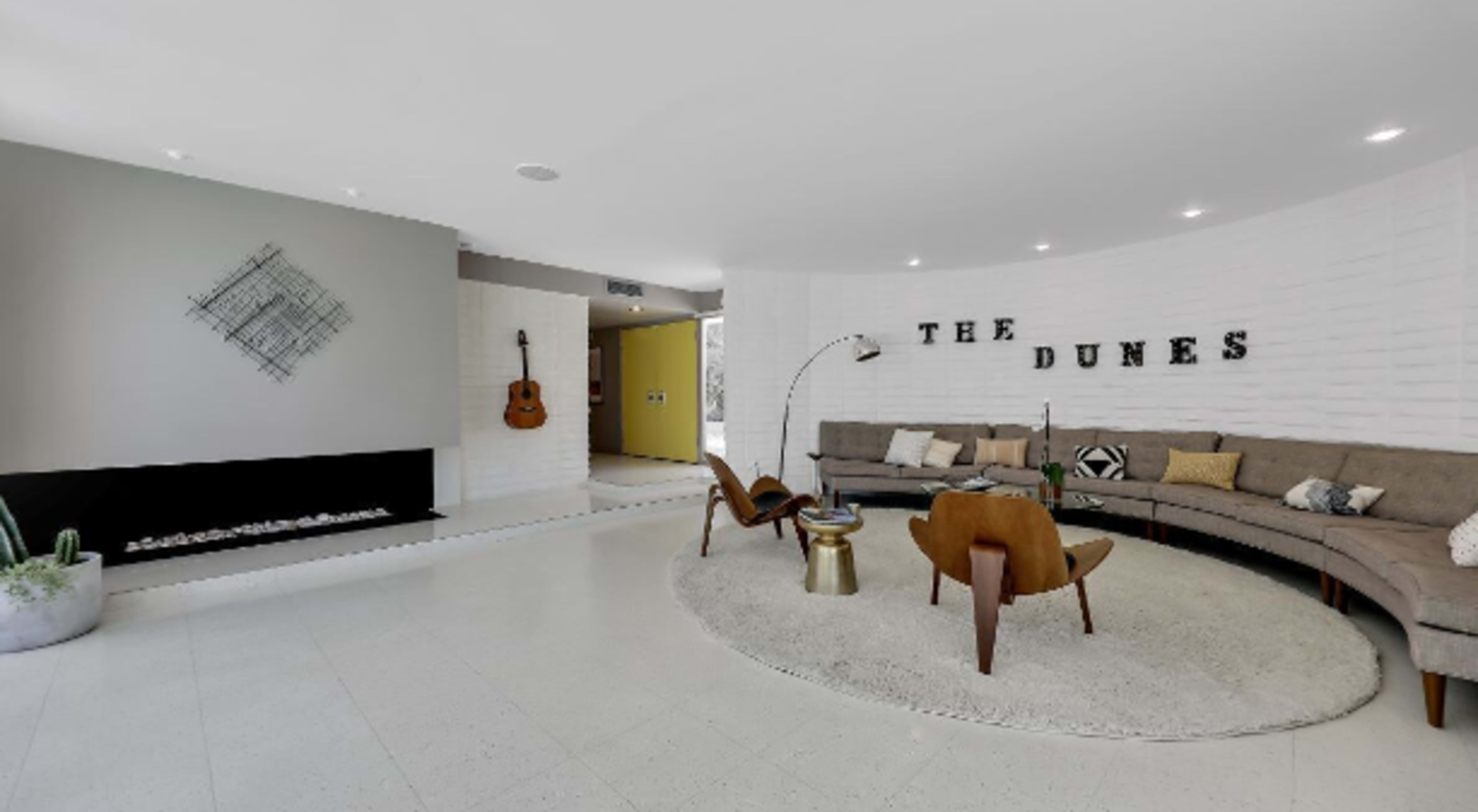 The image shows a modern living room with a curved sectional sofa, a round rug, a coffee table, and a fireplace, along with decorative wall text reading "THE DUNES."