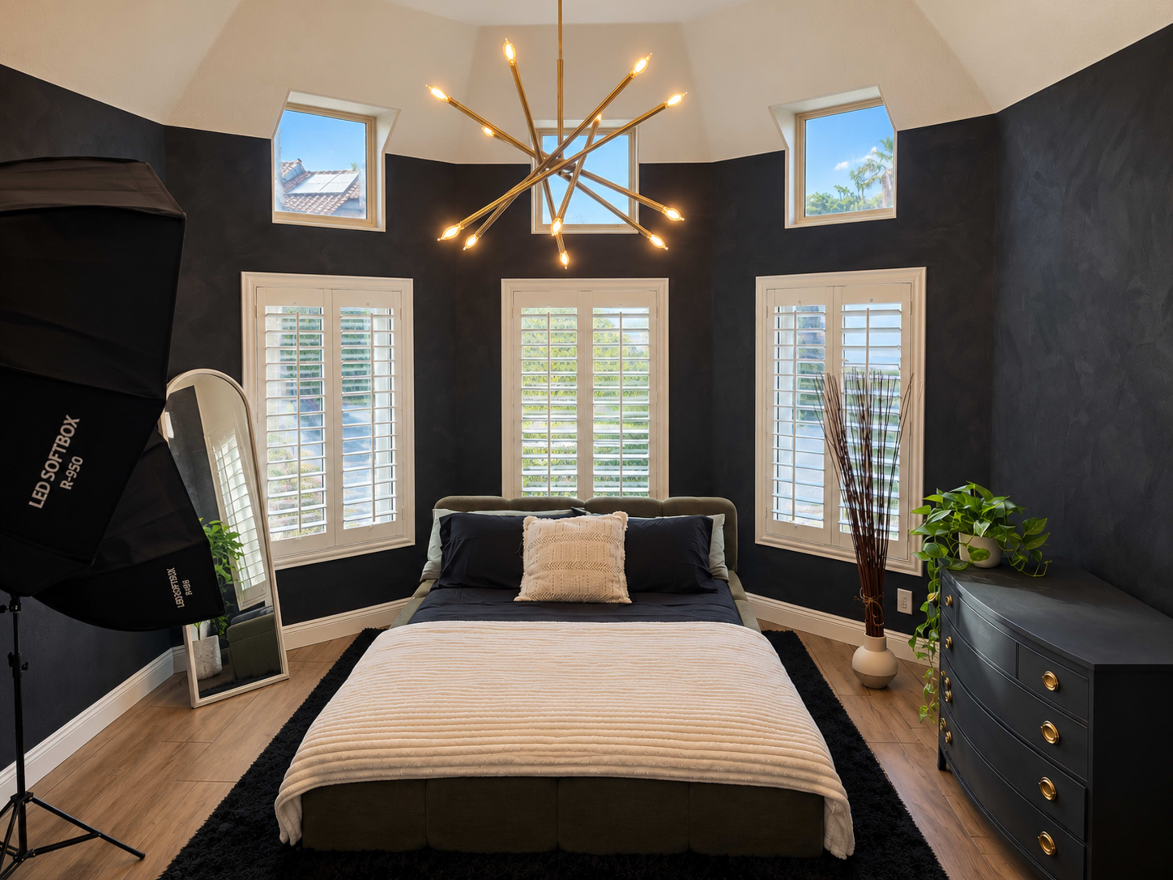 The bedroom features a bed with a layered blanket, modern lighting, large windows with plantation shutters, a dressing mirror, and a dark dresser.
