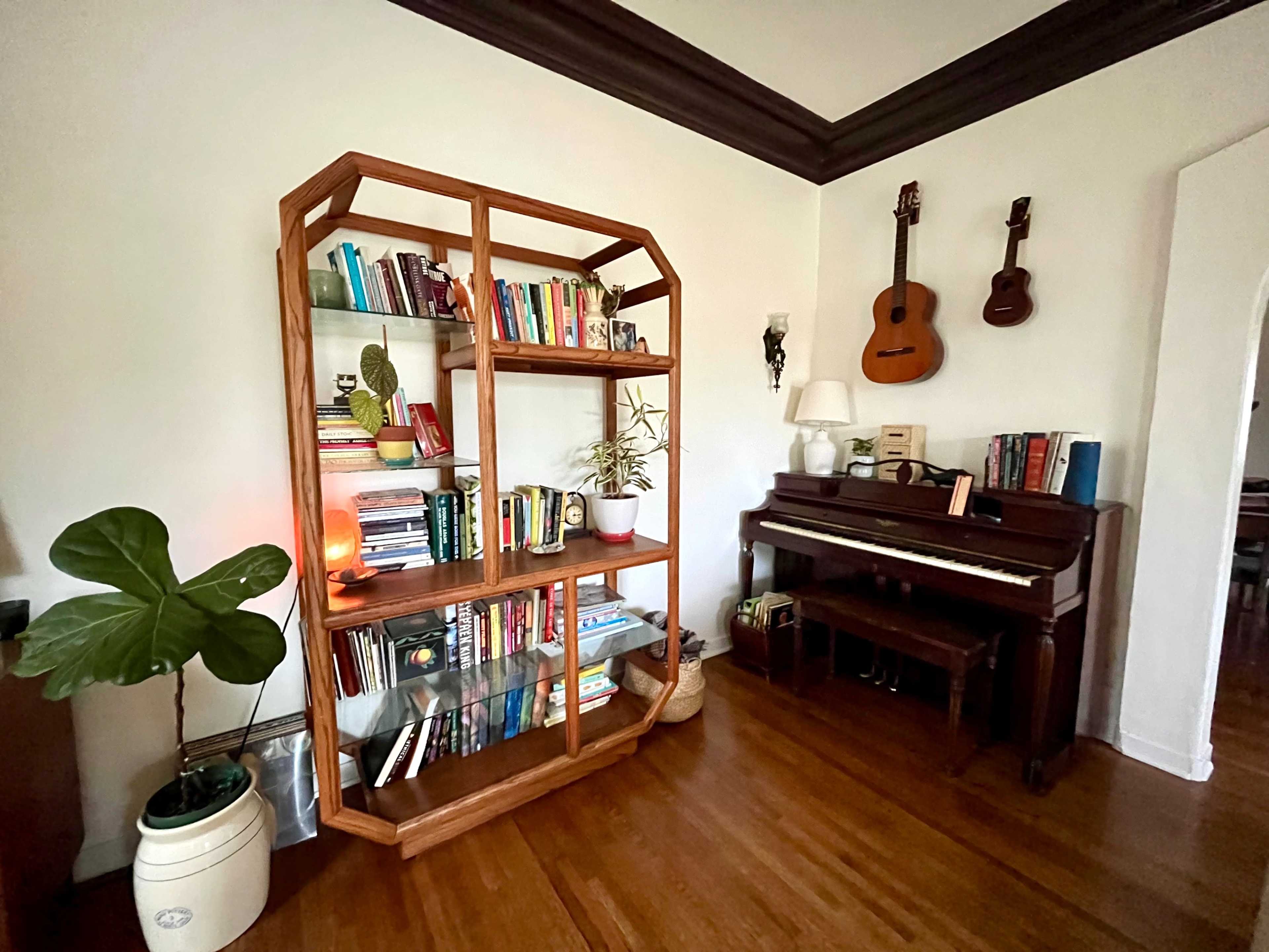 The image shows a cozy room featuring a wooden bookshelf filled with books, a piano, and a couple of guitars hanging on the wall.