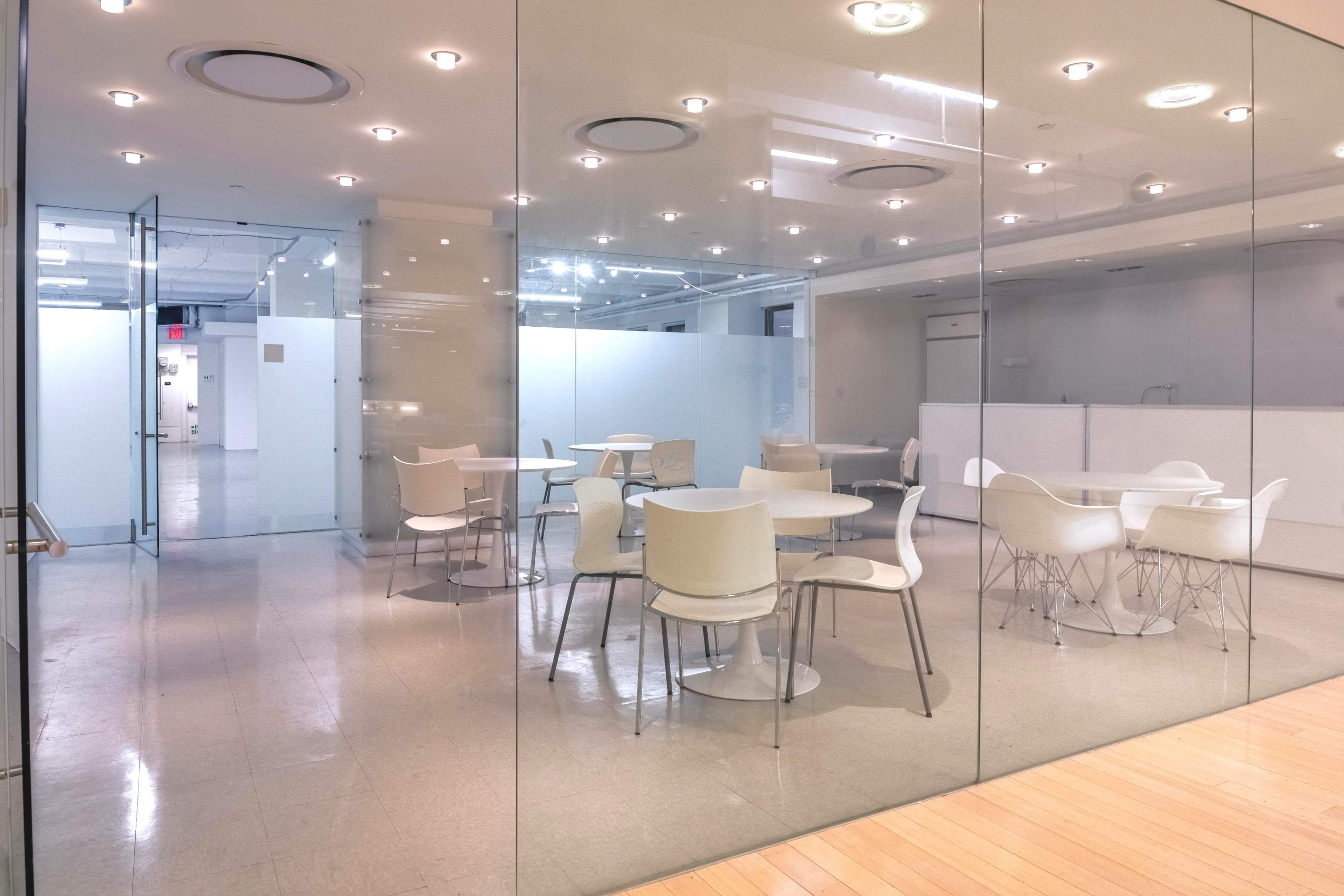 The image shows a modern, glass-walled office space with several round tables and white chairs arranged on a light-colored floor.
