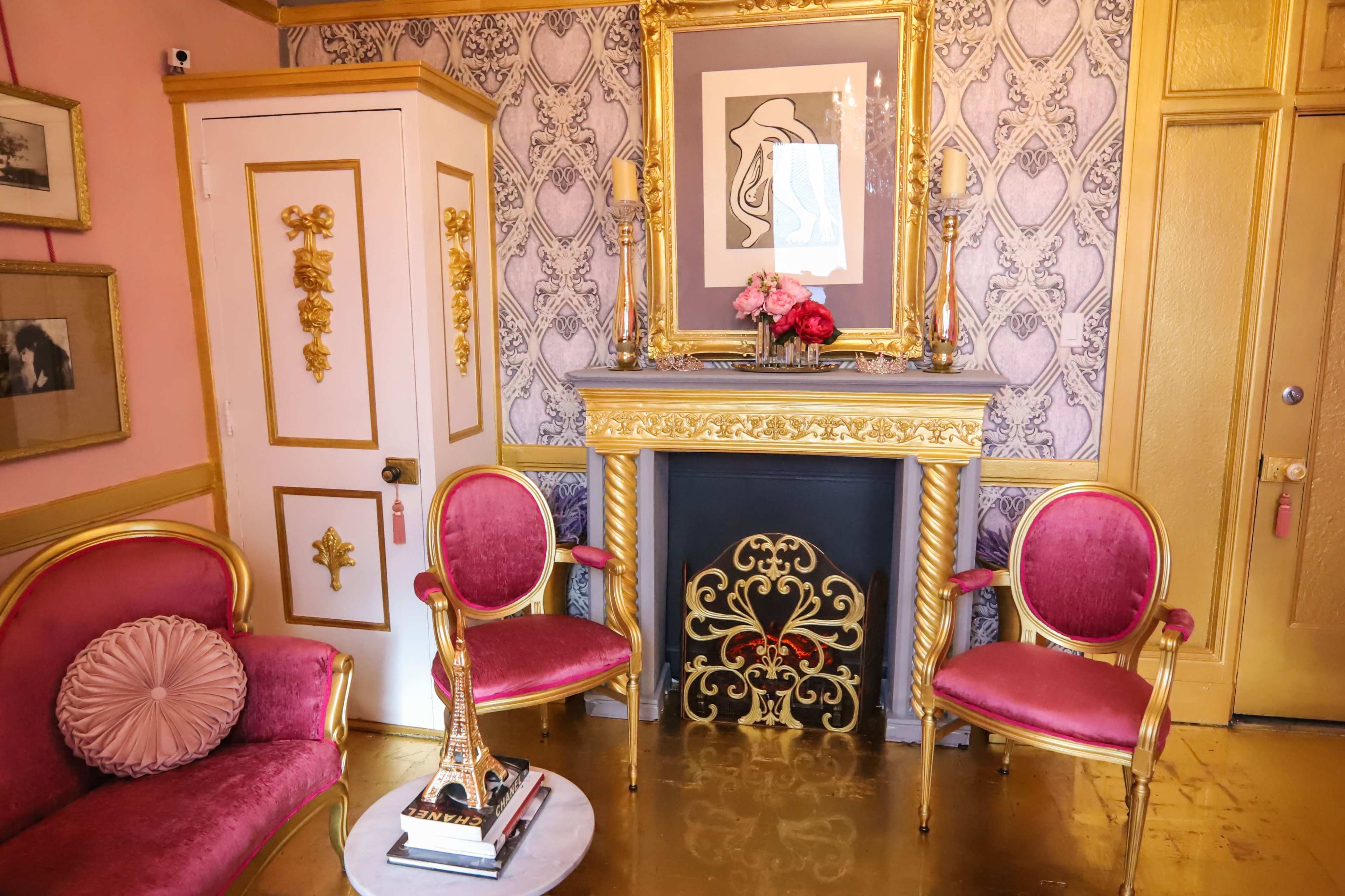 A decorated room featuring a gold-trimmed fireplace, two pink velvet chairs, and a table with an Eiffel Tower ornament, all against a patterned wallpaper.