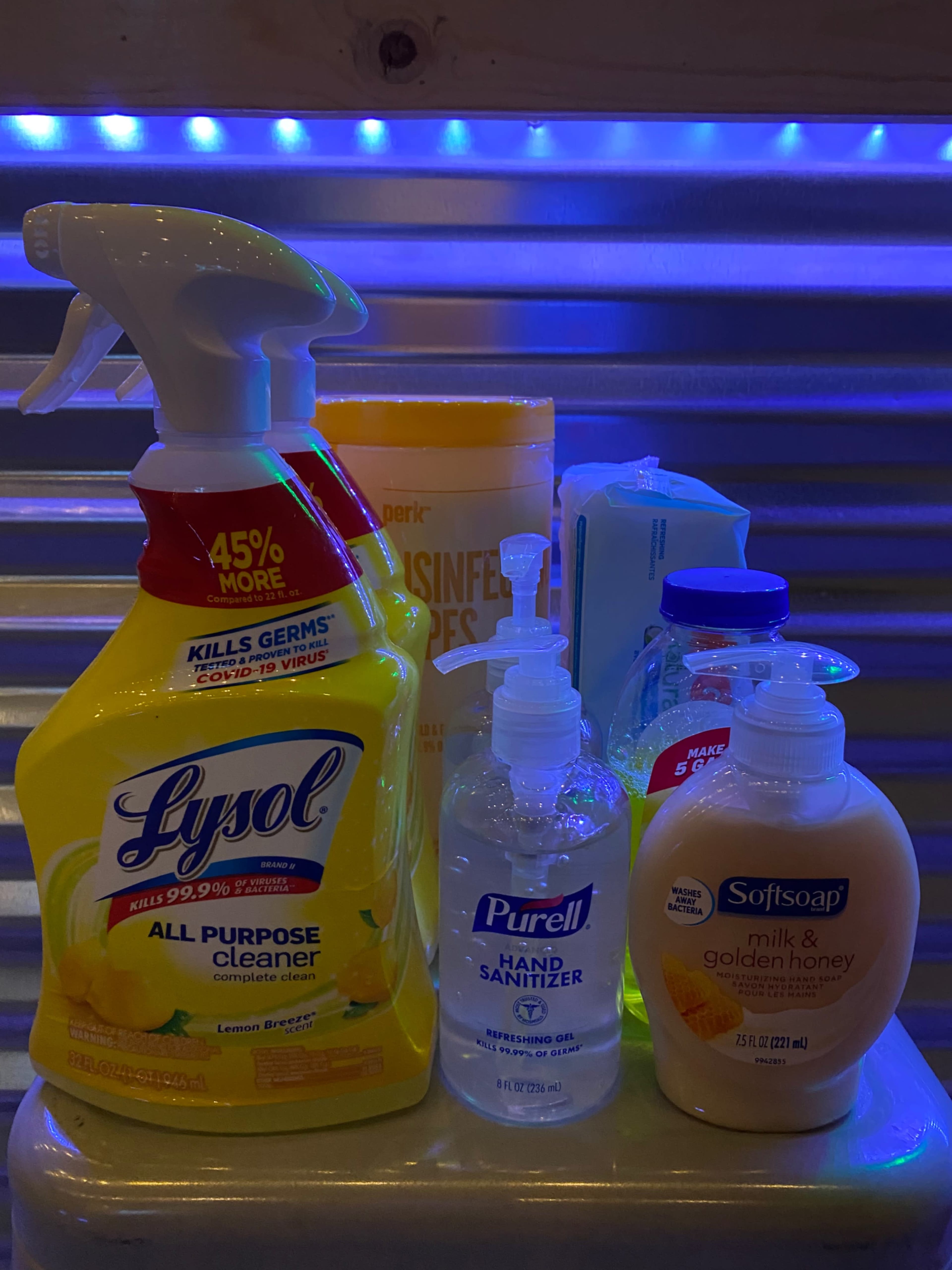 Various cleaning and sanitizing products, including a Lysol all-purpose cleaner, Purell hand sanitizer, disinfecting wipes, dish soap, and Softsoap hand soap, arranged on a surface with blue ambient lighting in the background.