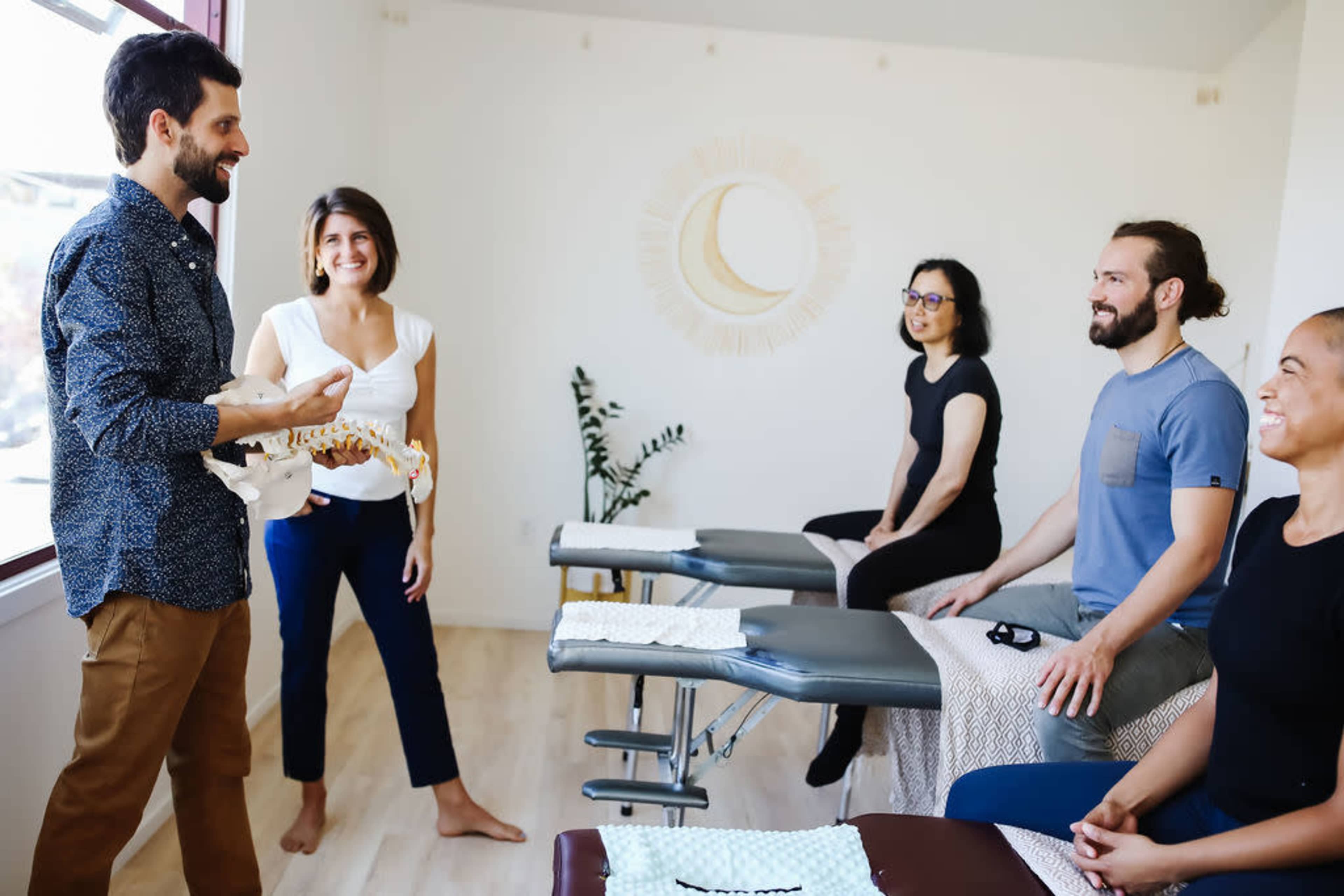 A therapist demonstrates a spine model to four seated individuals in a bright clinic setting.