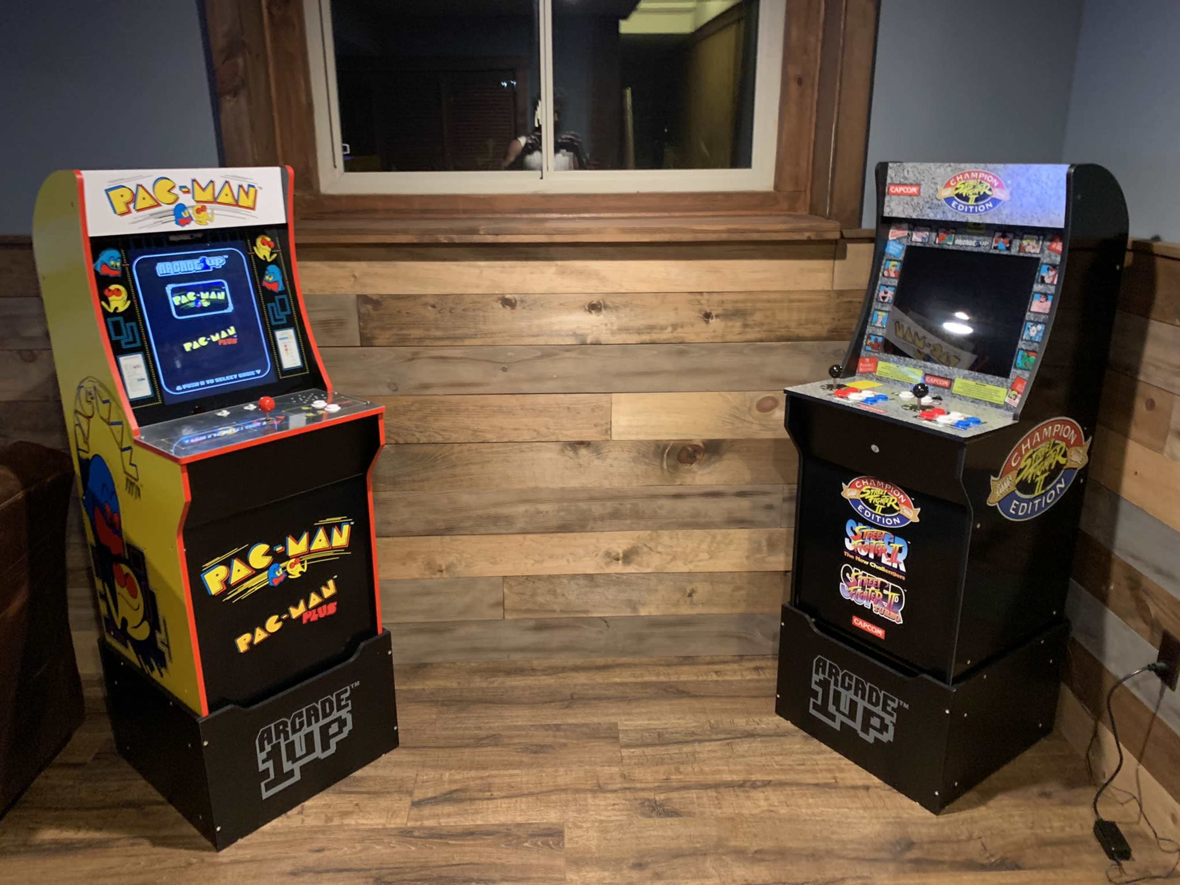 Two arcade game machines, one featuring Pac-Man on the left and the other labeled "Arcade 1UP" on the right, set against a wooden wall in a room.