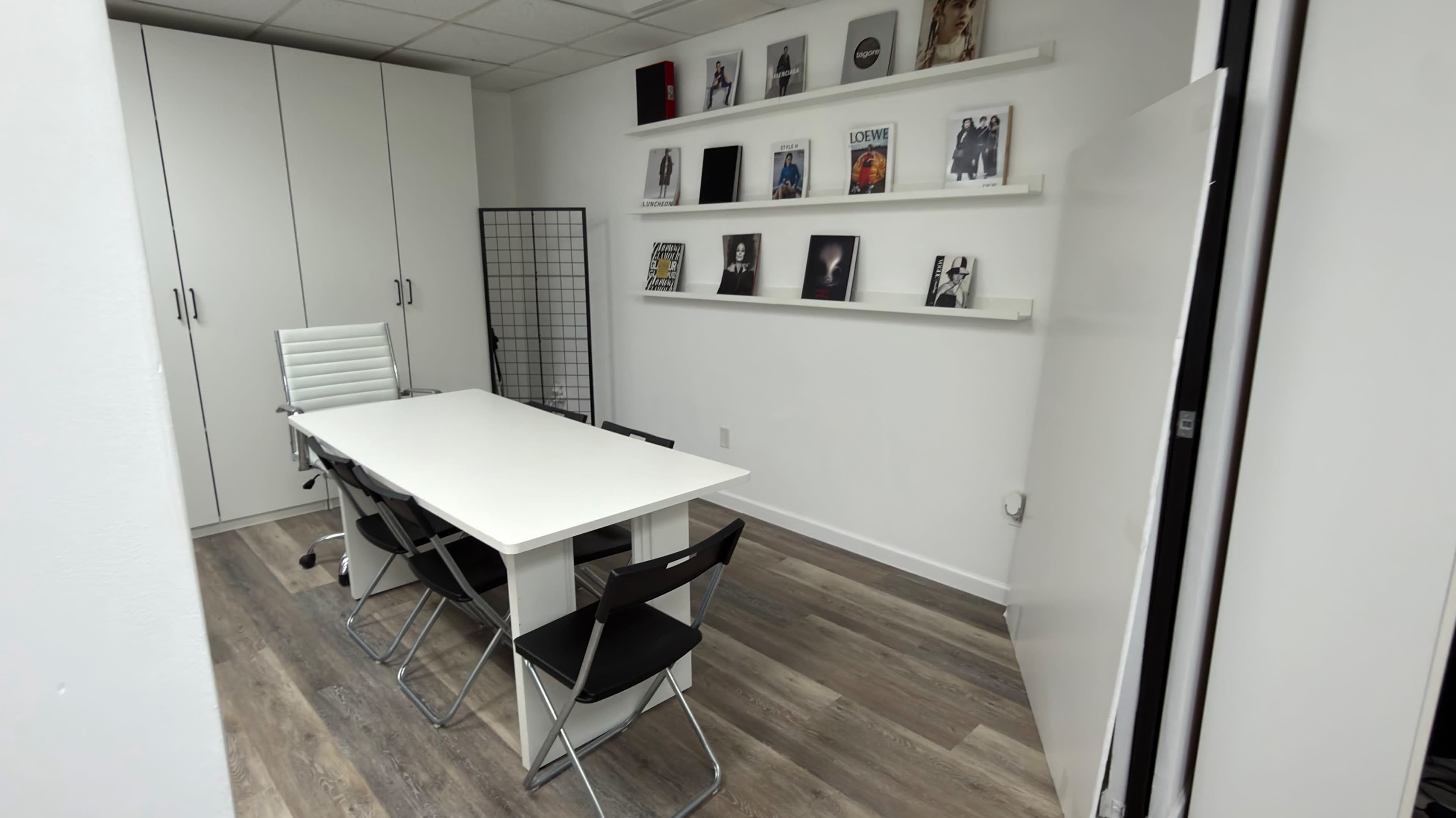 The image shows a modern meeting room with a white table, several black and white chairs, and a shelf displaying framed pictures on the wall.