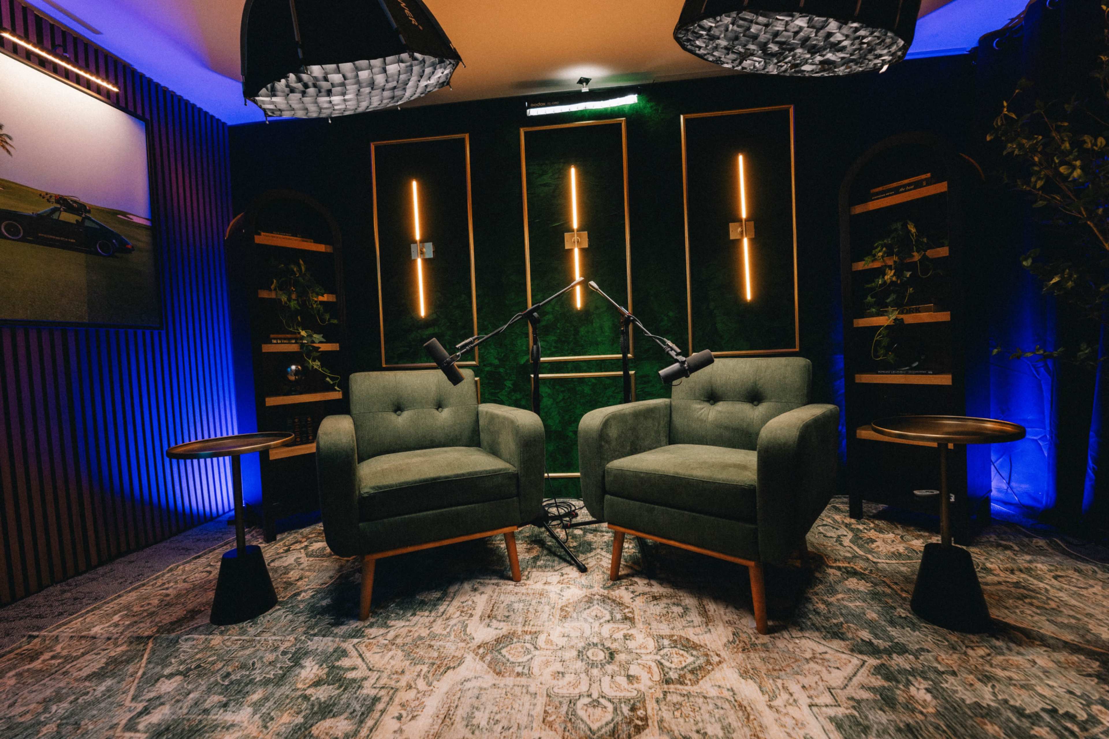 The image shows a cozy interview setup featuring two green armchairs, two small side tables, and ambient lighting on a patterned rug against a dark background.