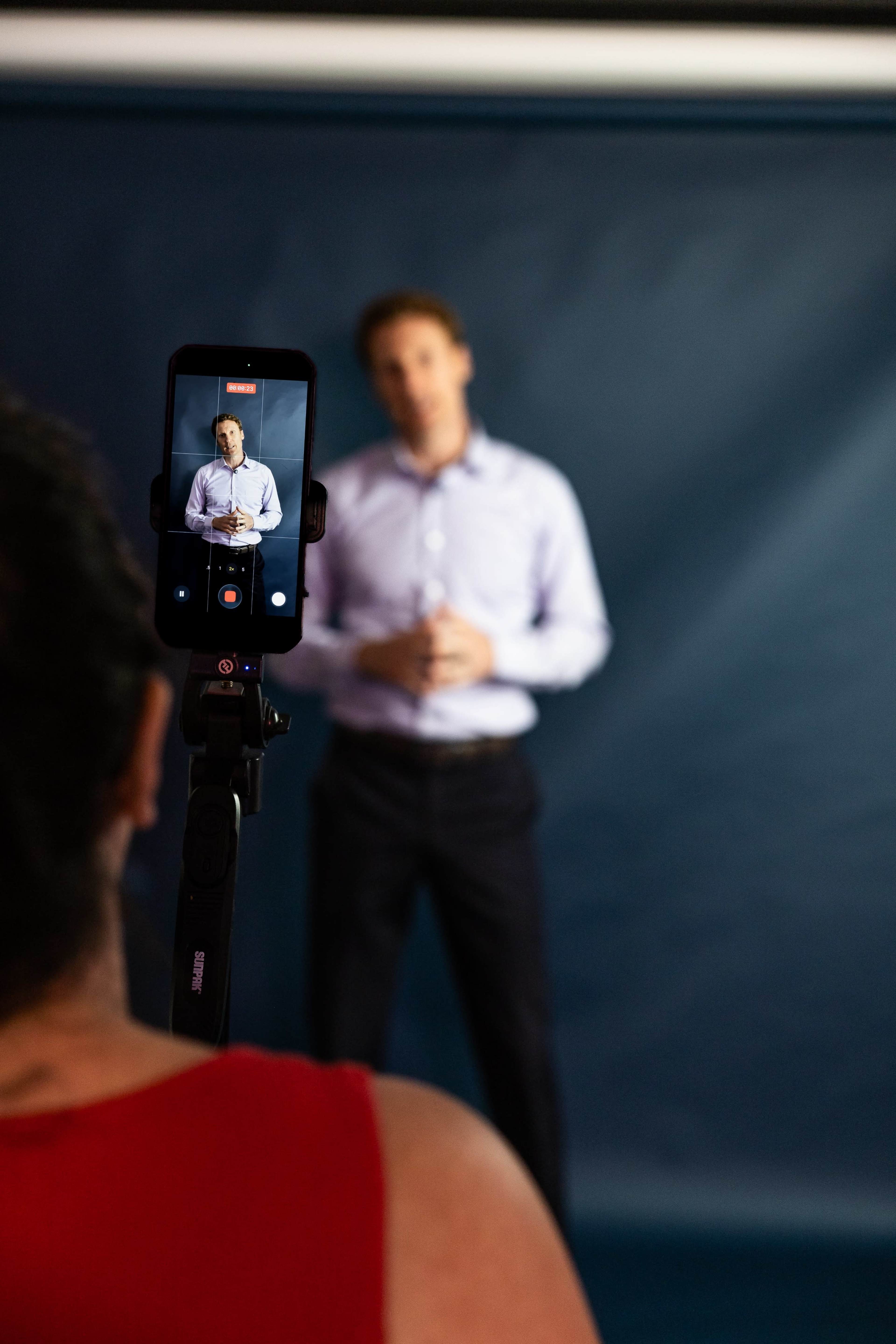 A person is filming another individual who is standing in front of a blue backdrop.