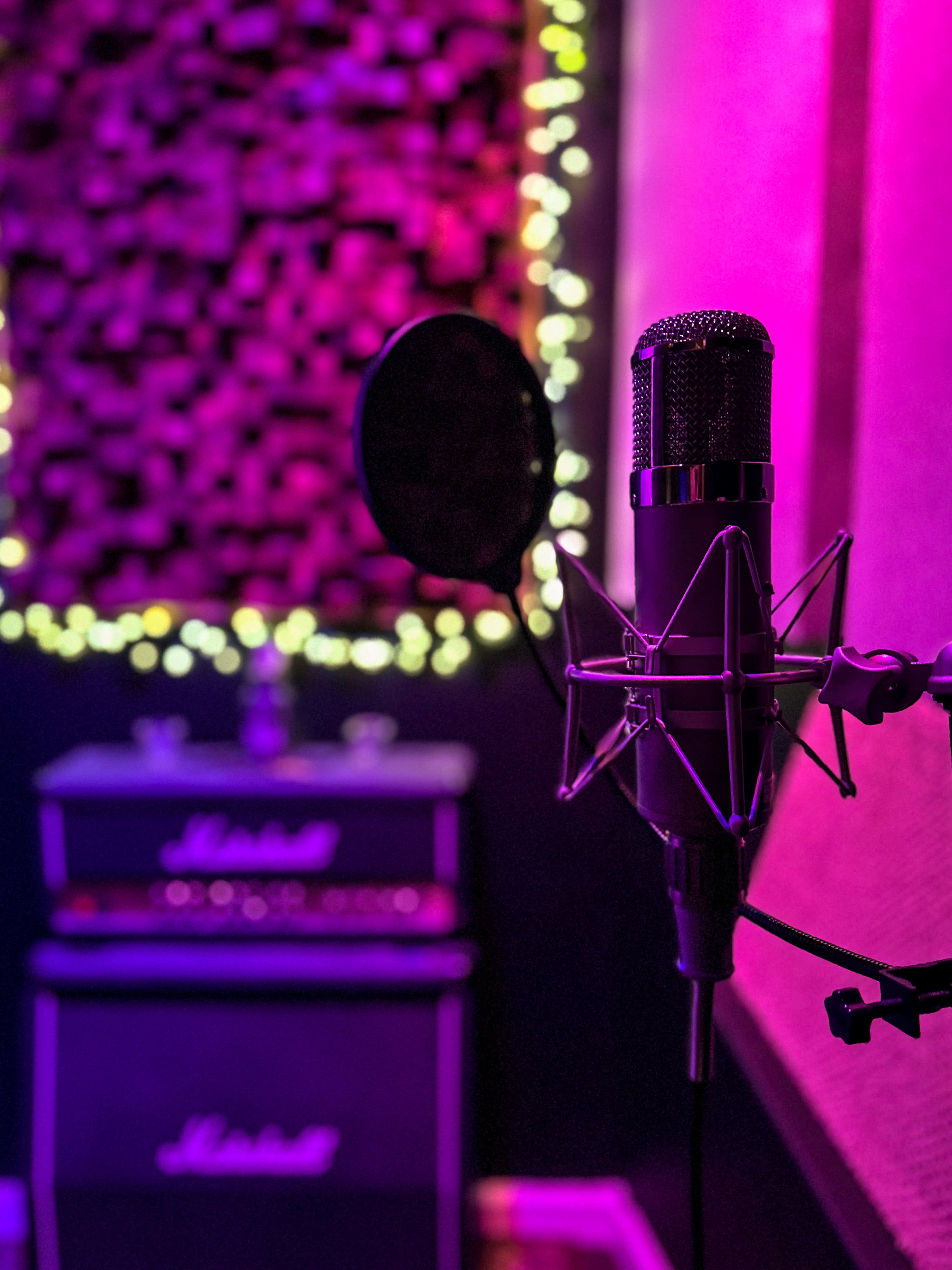 A microphone stands in the foreground of a recording studio, with a Marshall amplifier and soft lighting in the background.