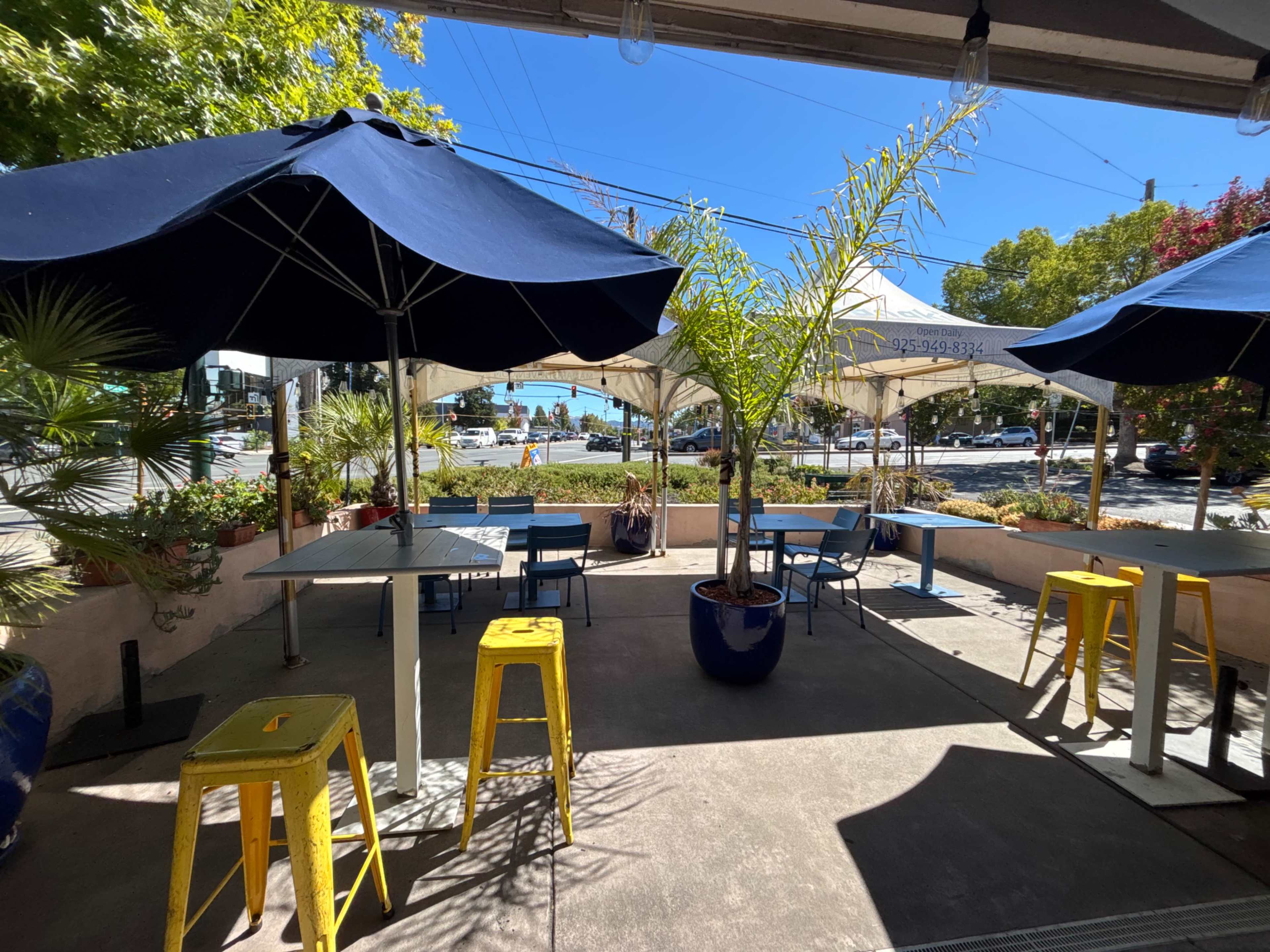 The image shows an outdoor dining area with blue umbrellas, yellow bar stools, and potted plants, overlooking a street.