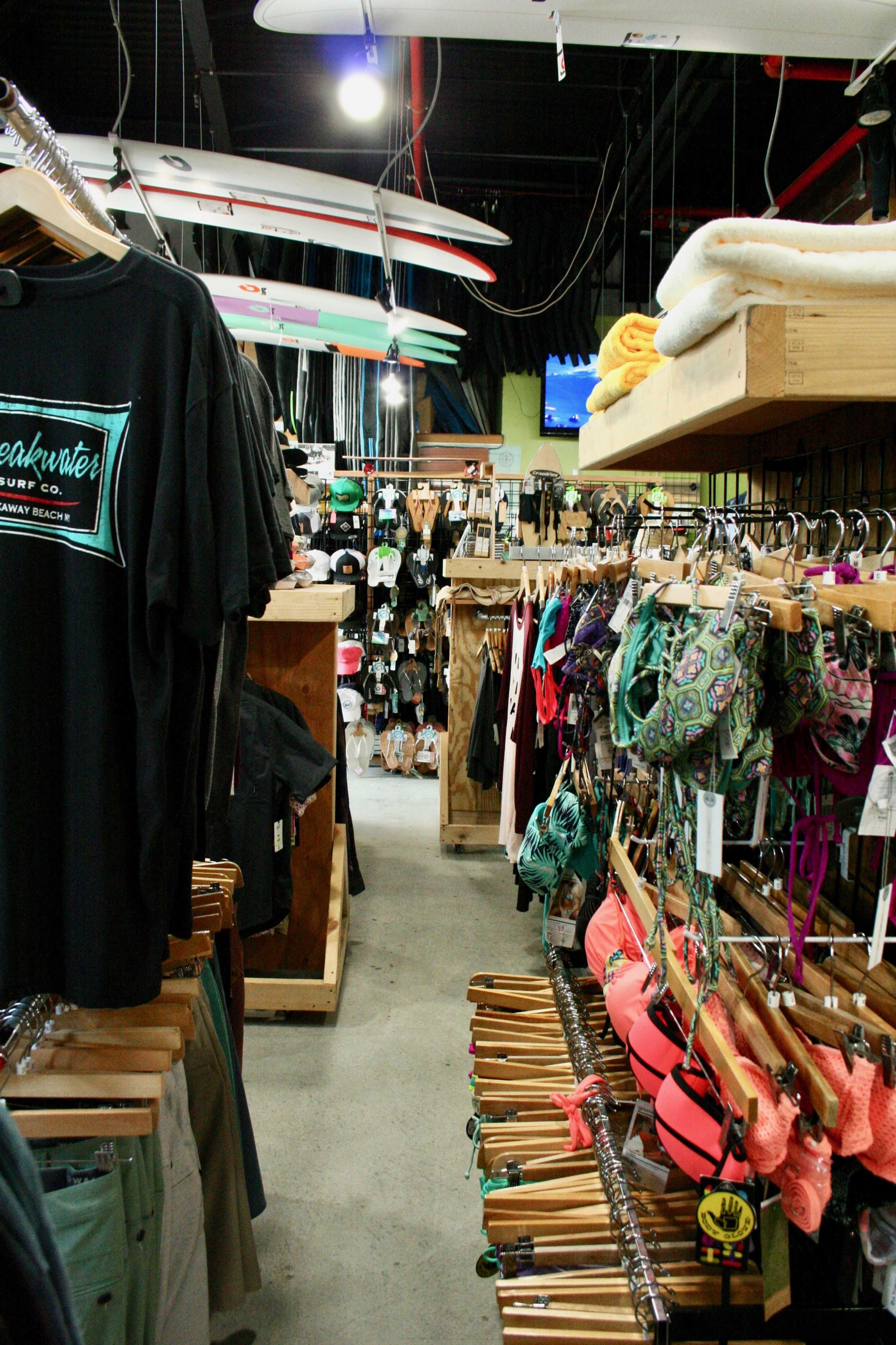 A narrow aisle in a surf shop displays rows of clothing and swimwear, with surfboards hanging overhead.