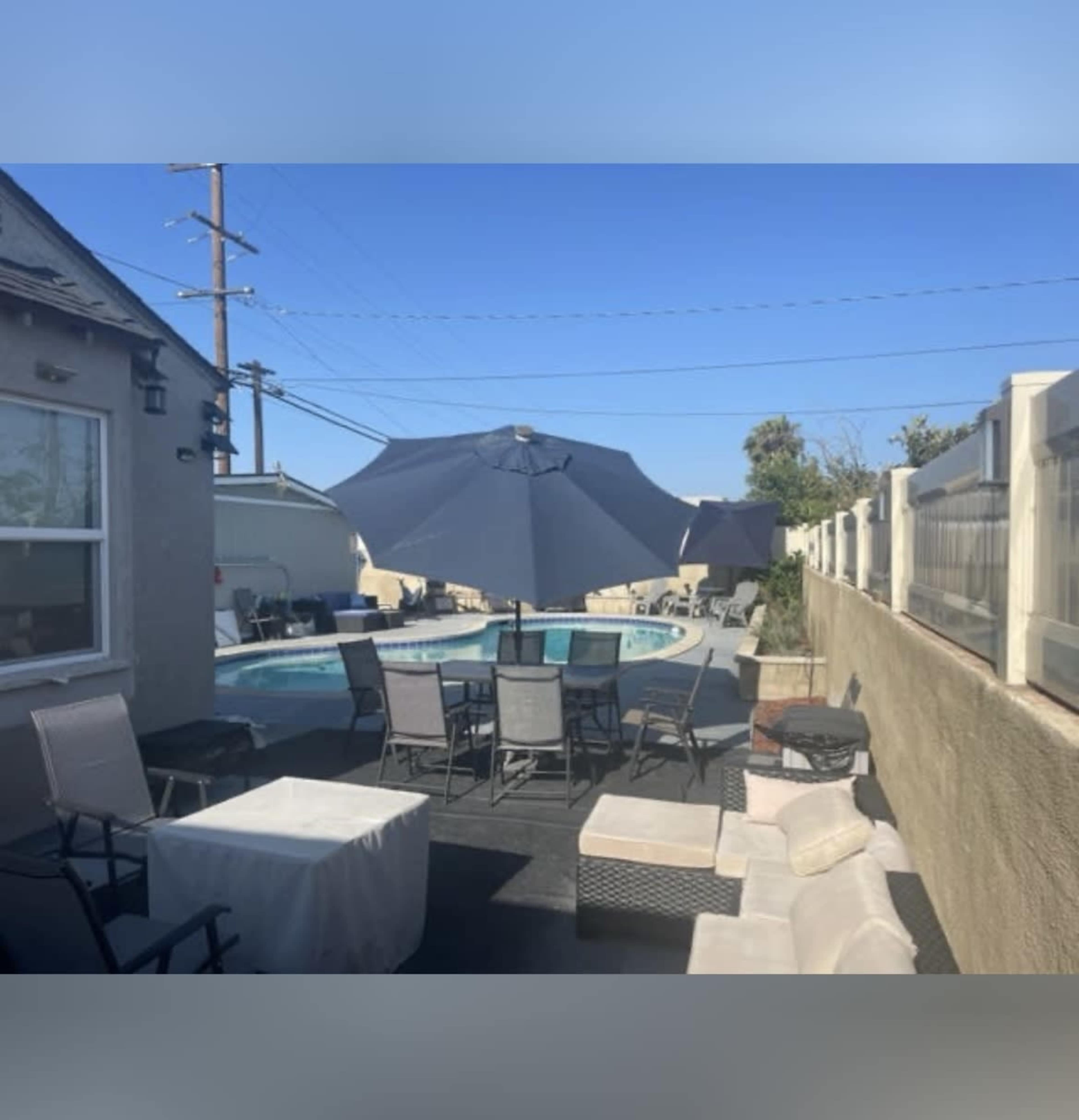 A pool area is visible with lounging chairs, tables, and large umbrellas, surrounded by a fence and a clear blue sky.