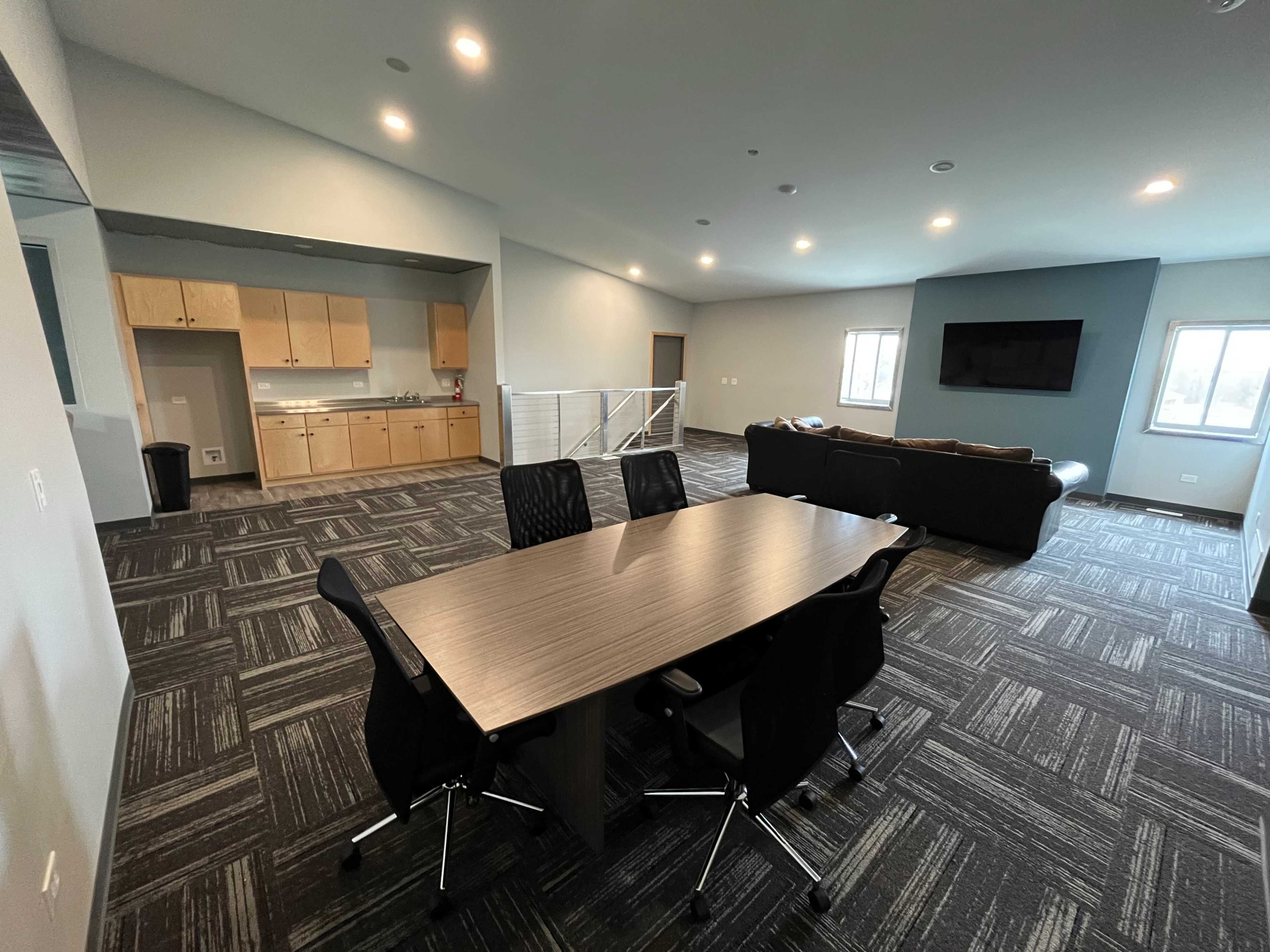 The image shows a spacious, modern office area with a large conference table, black chairs, a kitchenette, and a seating area featuring a couch and a television.