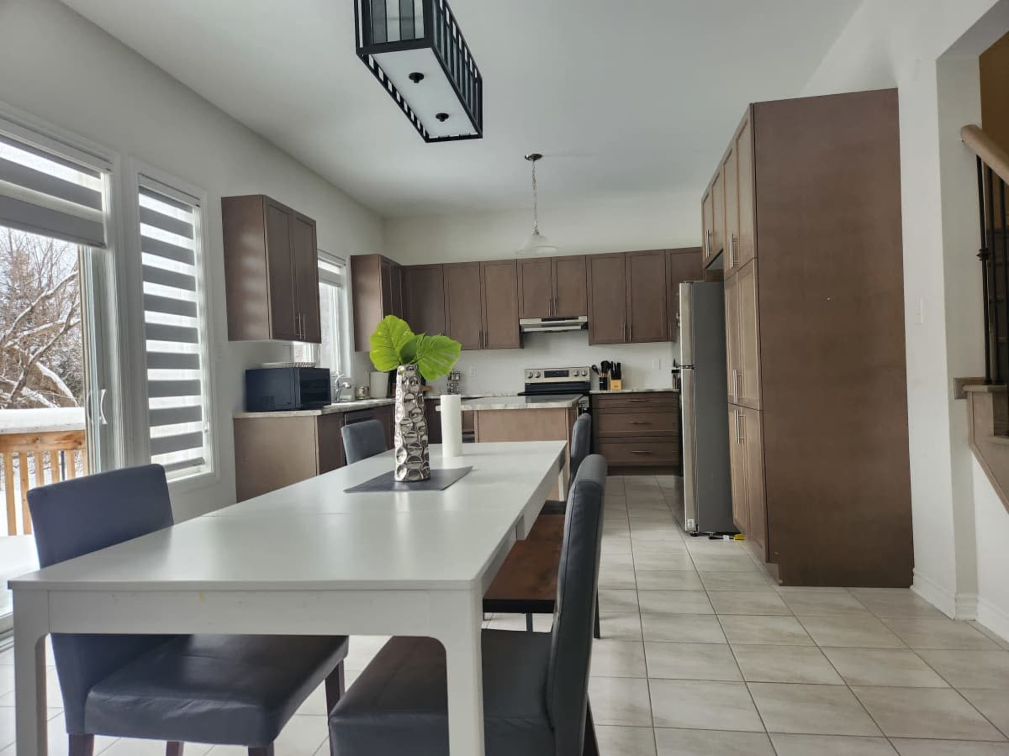The image shows a modern kitchen with wooden cabinets, a dining table surrounded by chairs, and large windows allowing natural light to enter.