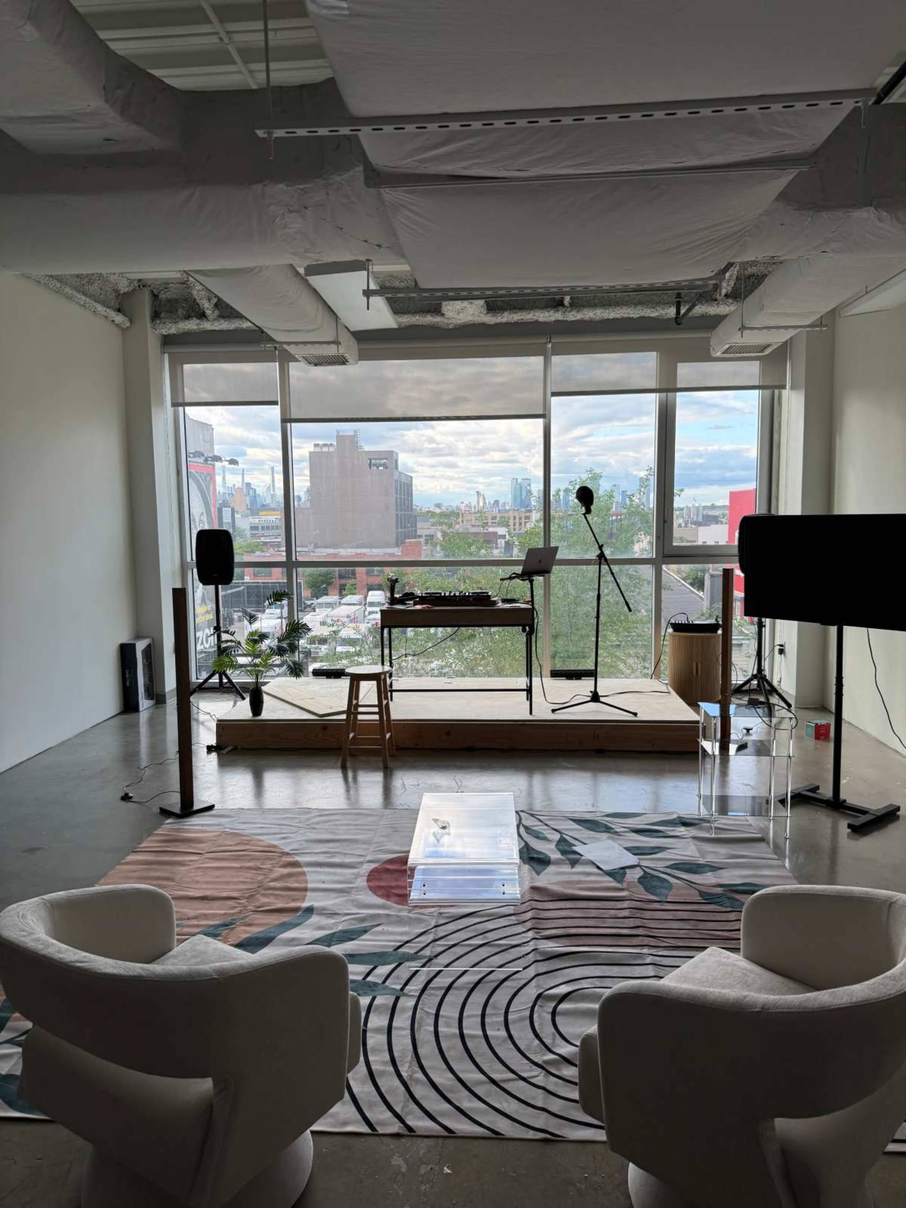 The image shows a modern office space with two white chairs, a large window overlooking a cityscape, and a wooden platform equipped with a keyboard and microphone setup.