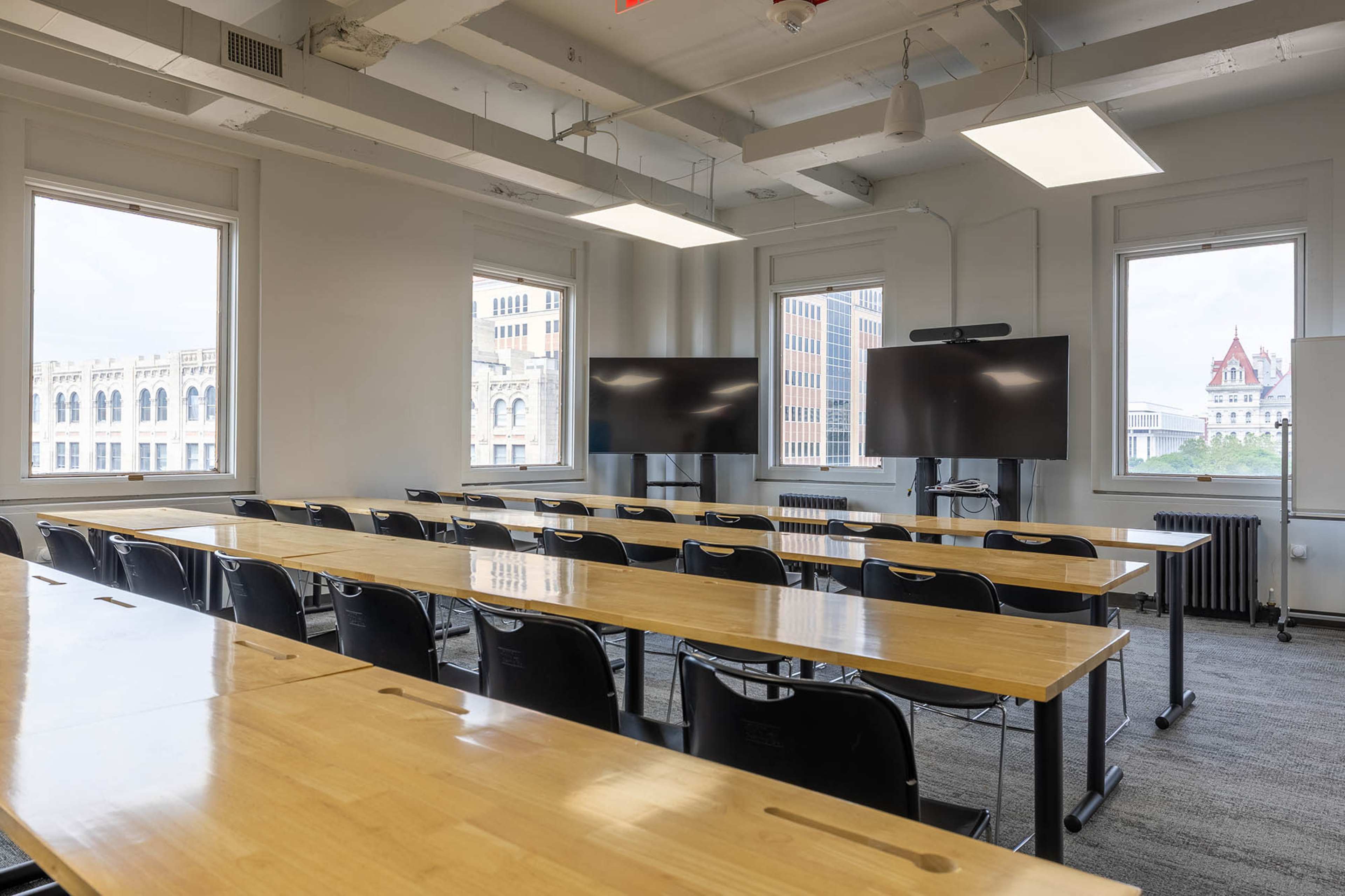 The image shows a modern classroom with rows of wooden tables and black chairs, featuring two large screens at the front and windows providing natural light.