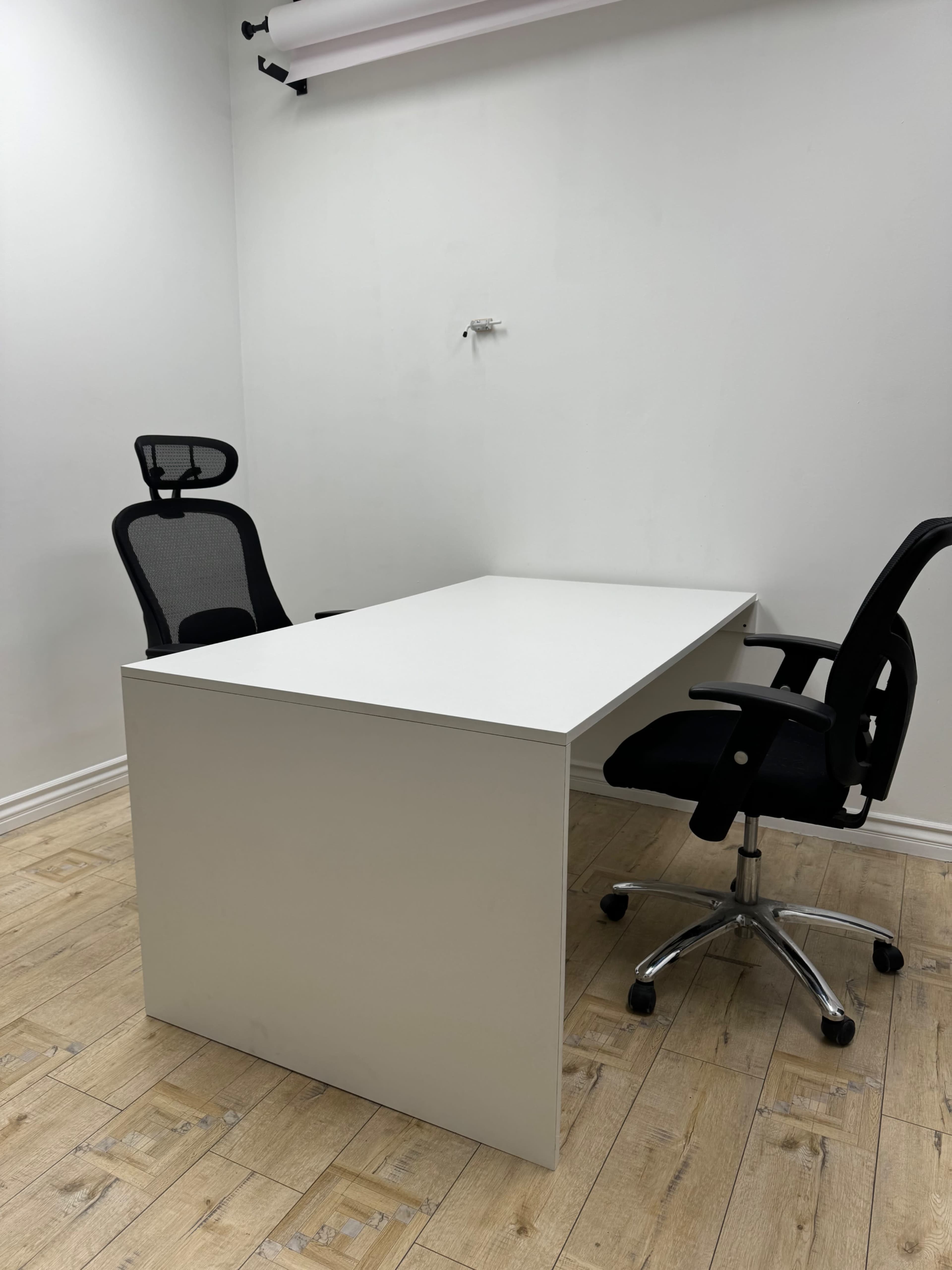 A simple office space features a white desk and two black ergonomic chairs against a plain white wall.