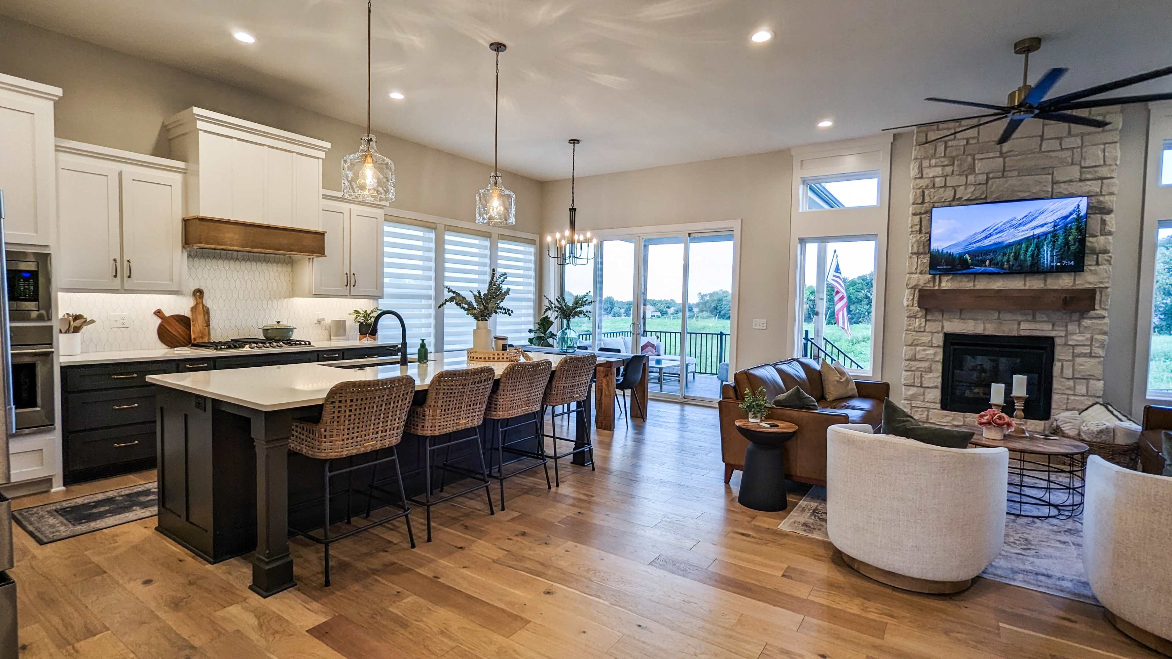 A modern open-concept kitchen and living area features a large island with bar stools, stone accents, and expansive windows overlooking a scenic view.