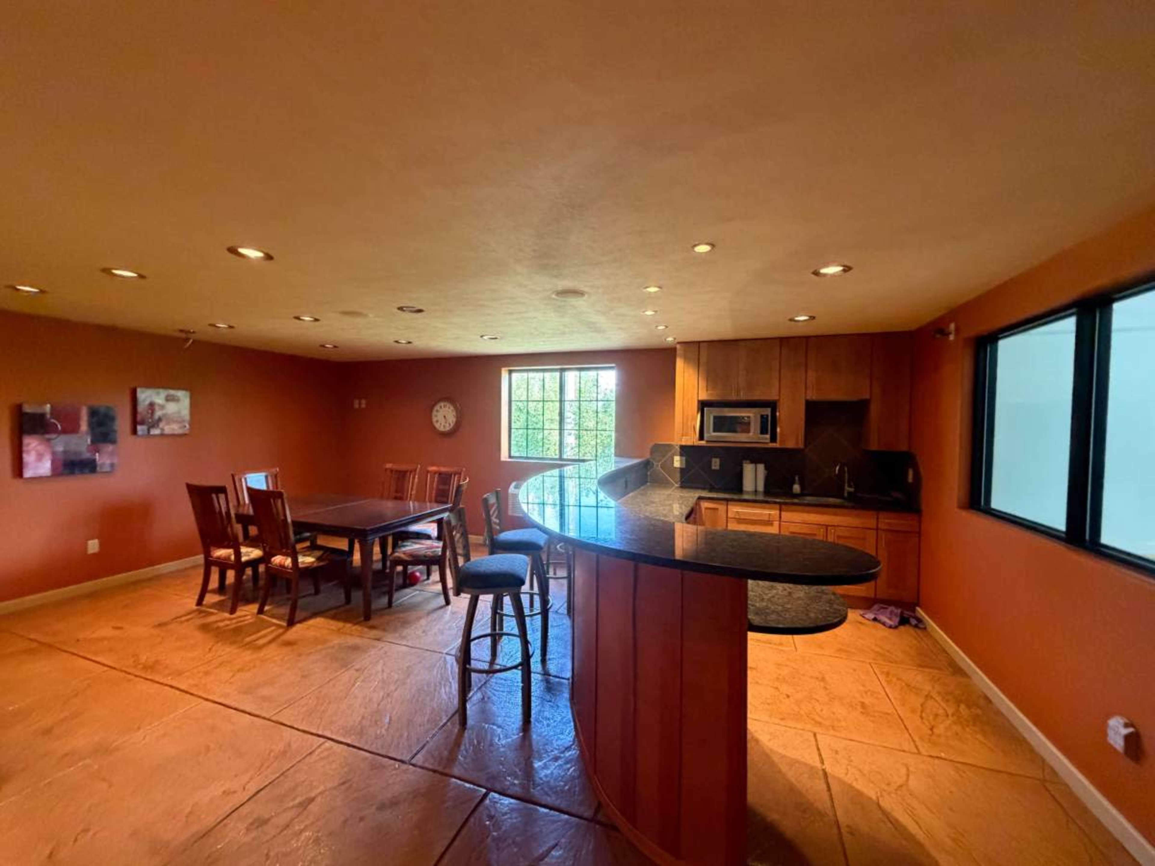 The image shows an open kitchen and dining area with a large table, bar-style seating, and warm-colored walls.