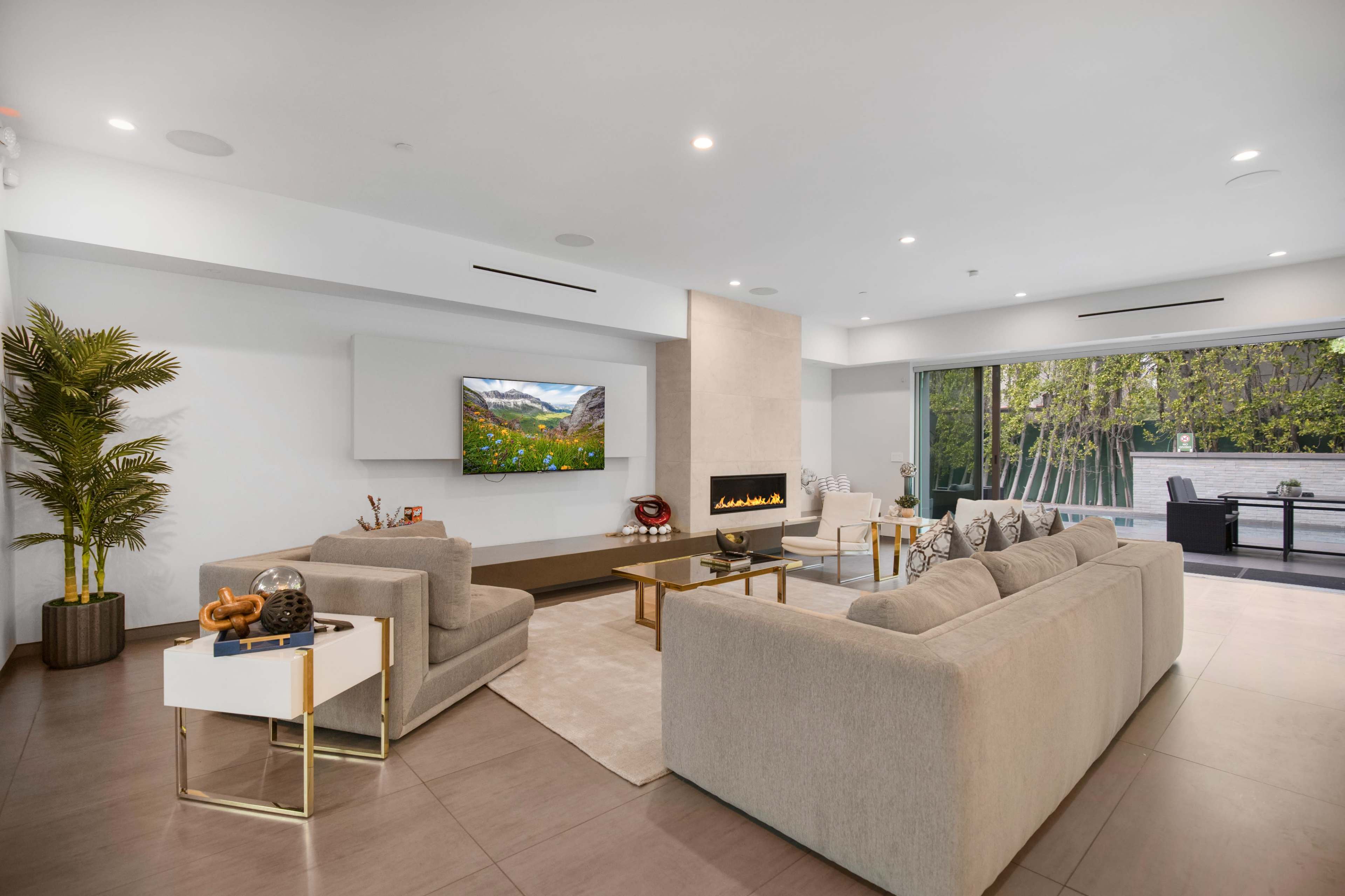 The image shows a modern living room featuring two light-colored sofas, a flat-screen TV mounted on the wall, and a large window that overlooks an outdoor space.