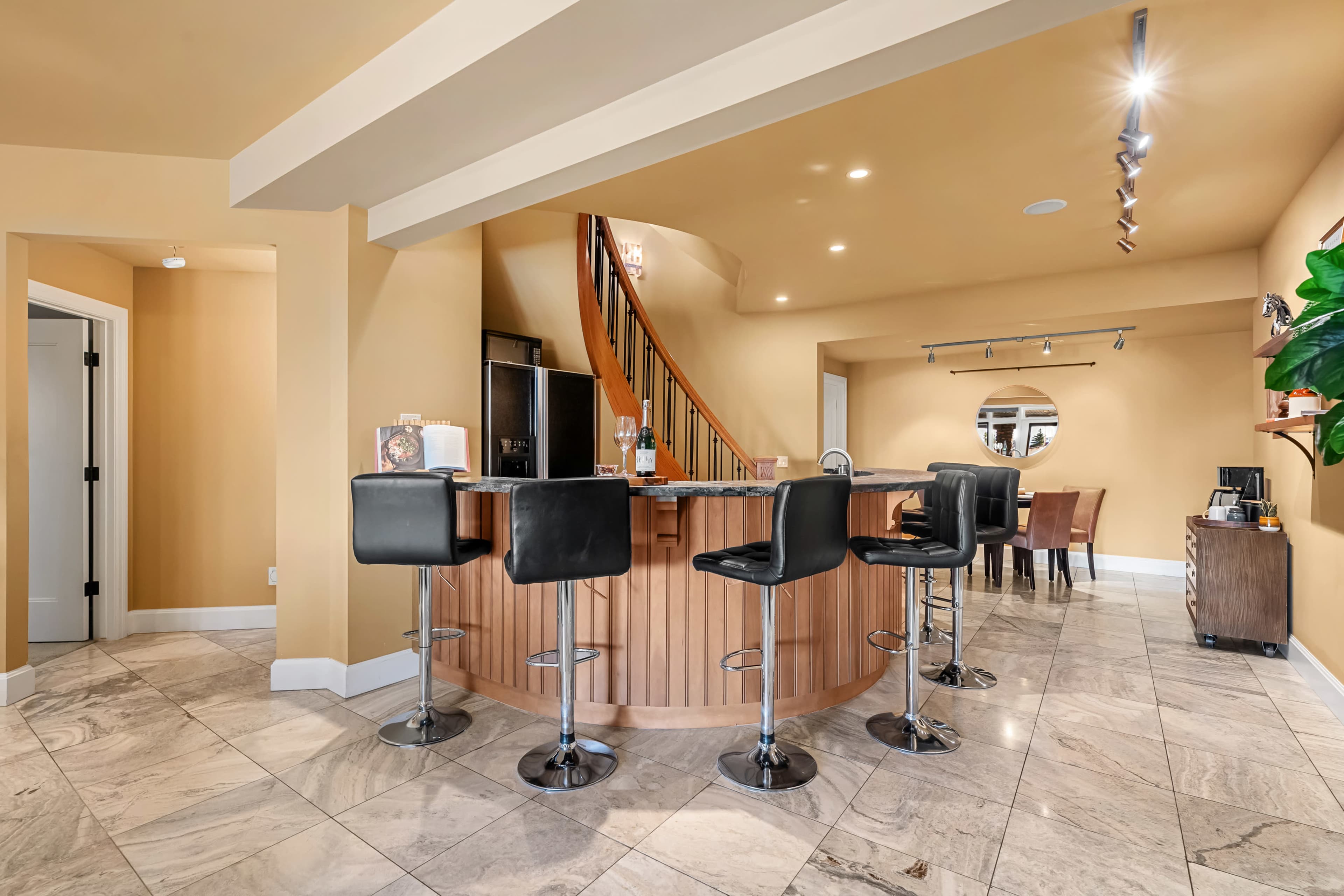 The image shows a modern home bar area with a circular wooden bar topped with four black stools, surrounded by beige walls and a staircase in the background.