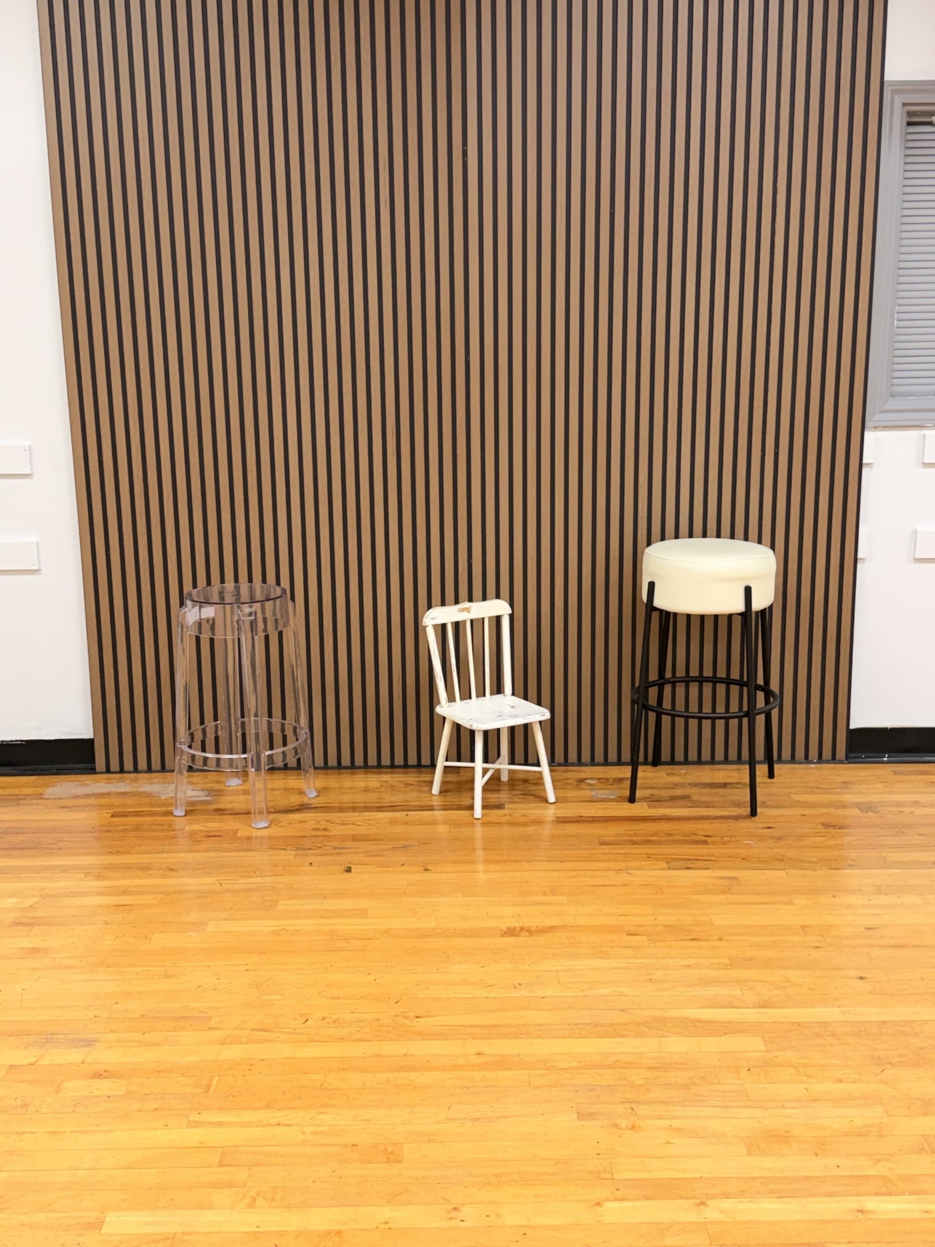 The image shows three chairs of different styles and colors lined up against a wooden slatted wall in a room with wooden flooring.