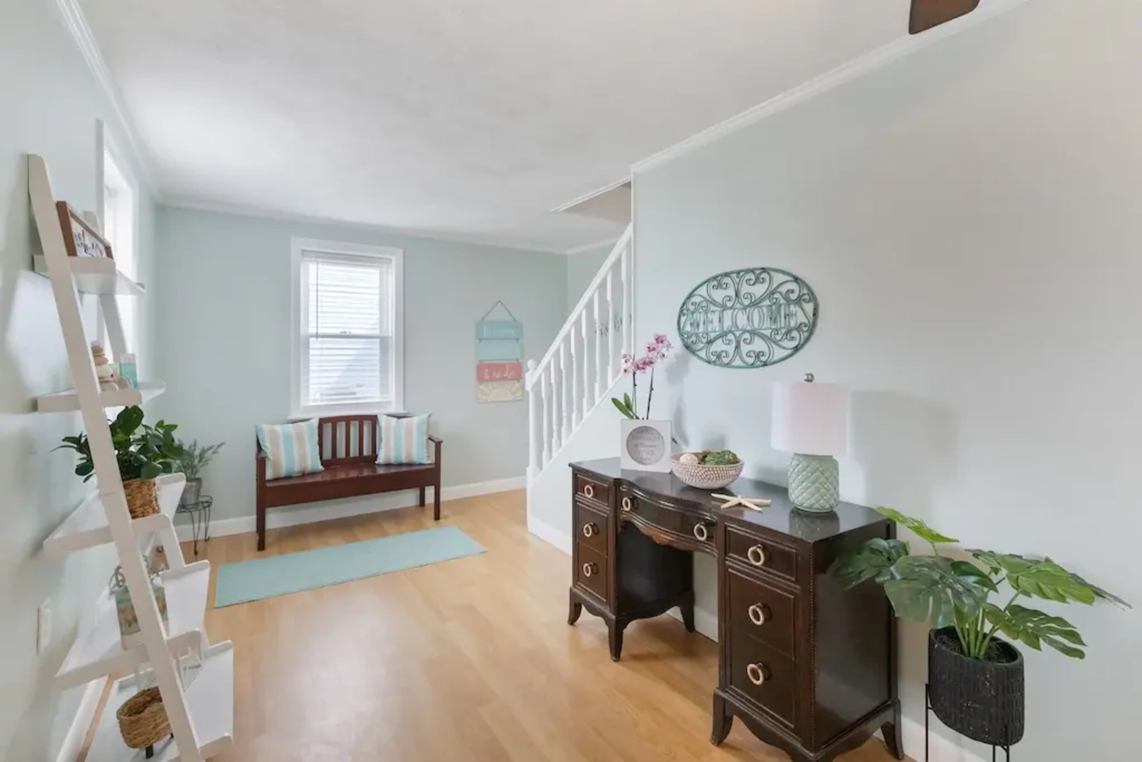 A bright, airy hallway with light blue walls, a wooden bench, a desk, a plant, and a staircase in the background.