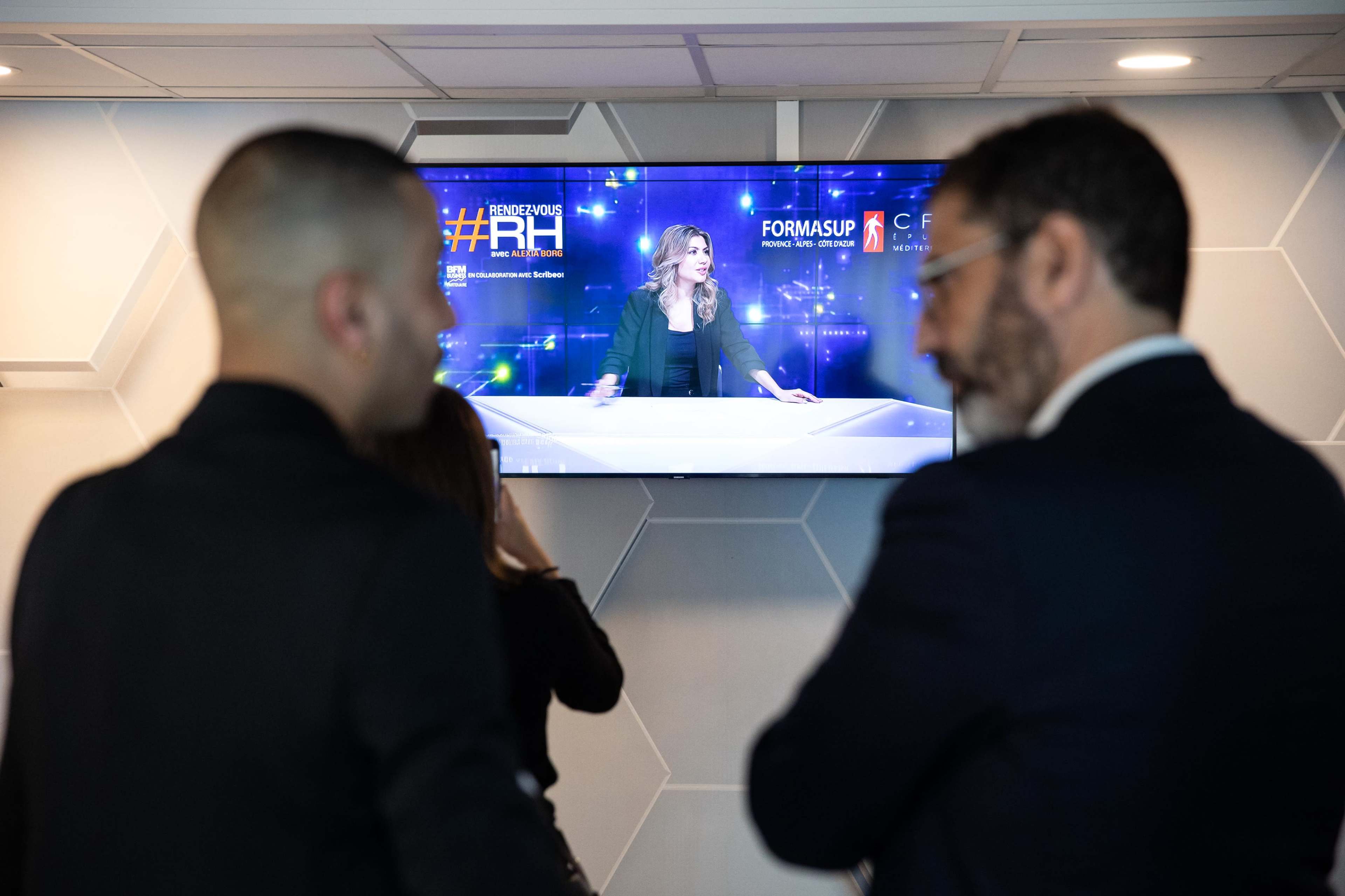Two people stand in front of a television displaying a news program or event hosted by a woman.