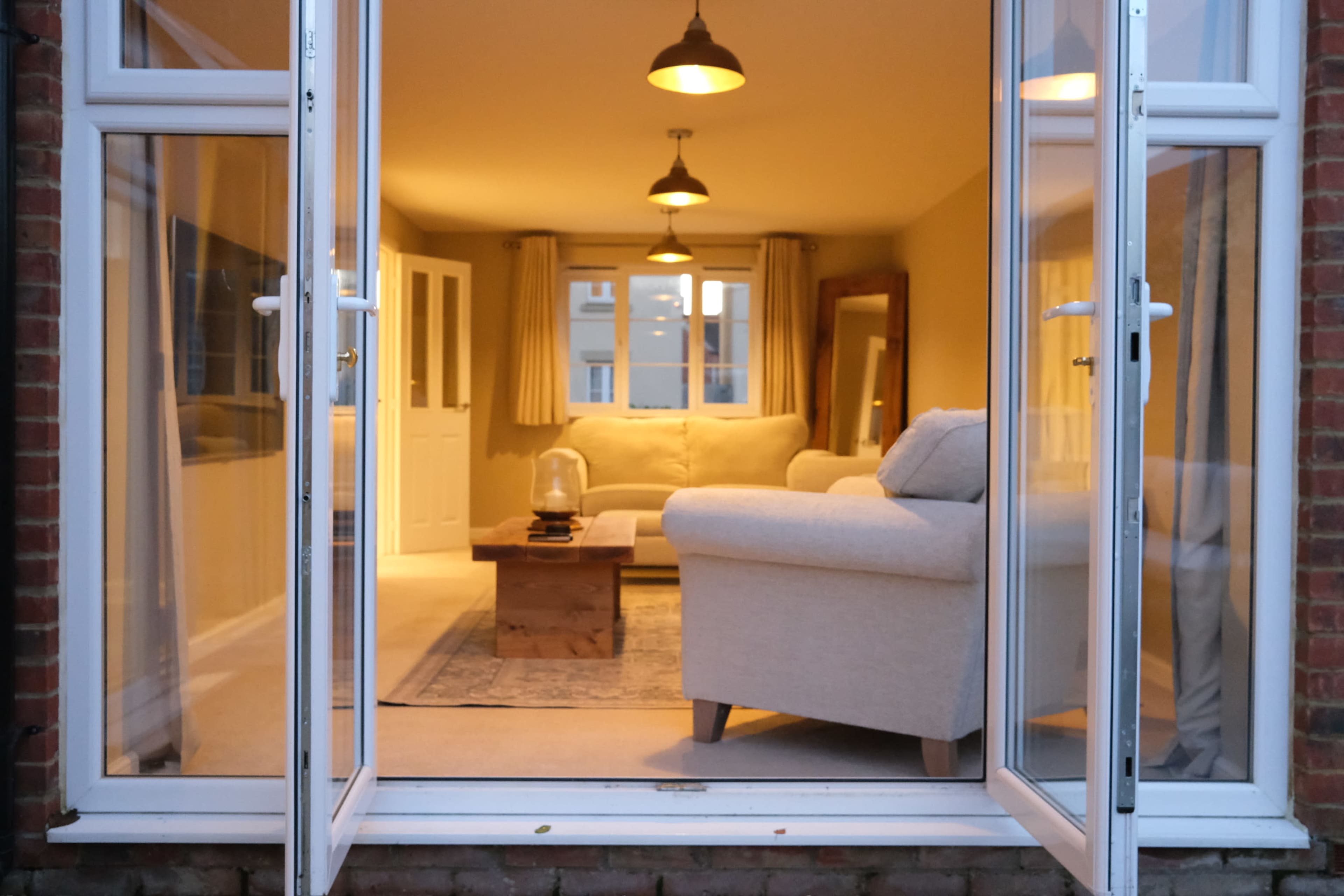 The image shows a cozy living room viewed through open French doors, featuring a light-colored sofa, wooden coffee table, and warm lighting.