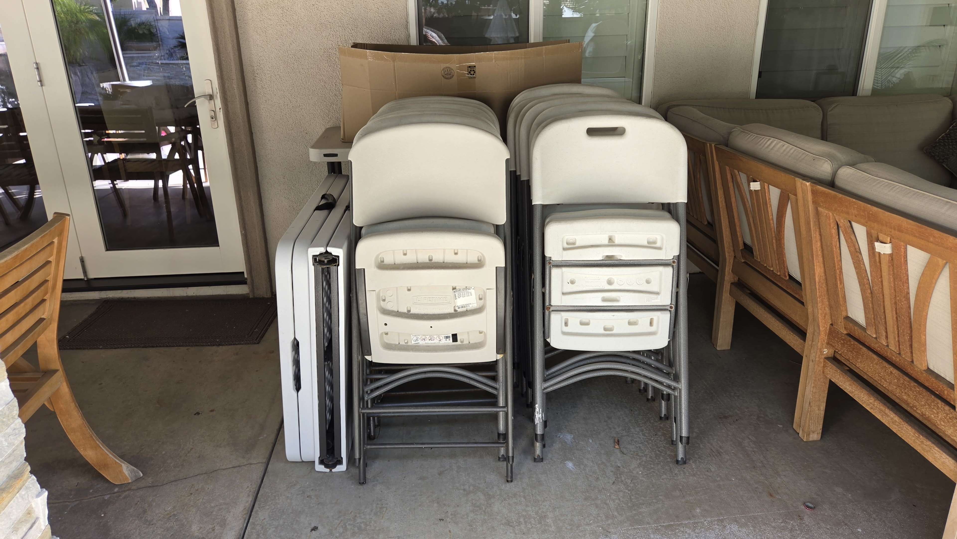 A stack of folded plastic chairs is placed next to a cardboard box and a couch on a patio.