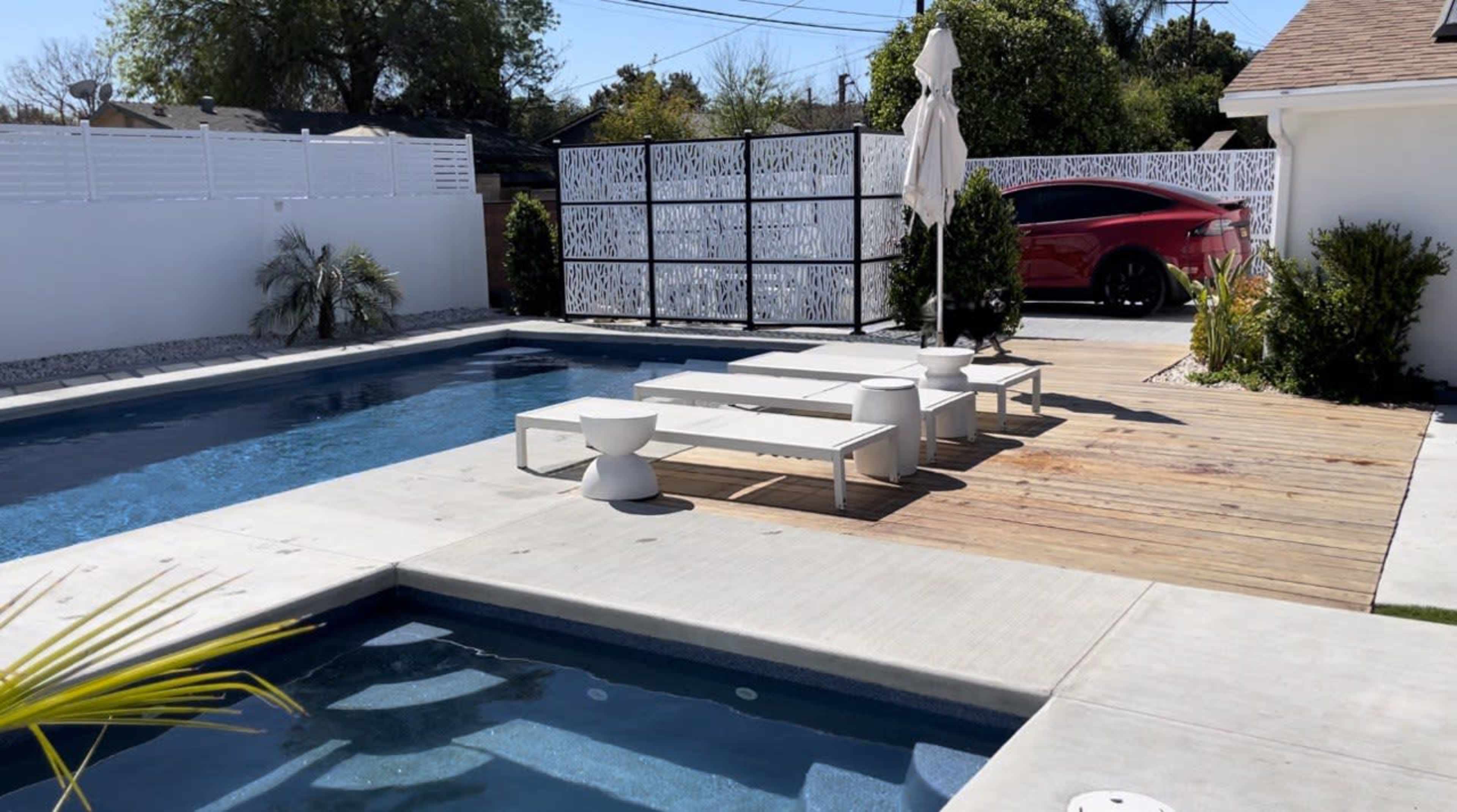 A modern backyard pool area featuring a large swimming pool, a wooden deck with white outdoor furniture, and a sleek car parked in the background.