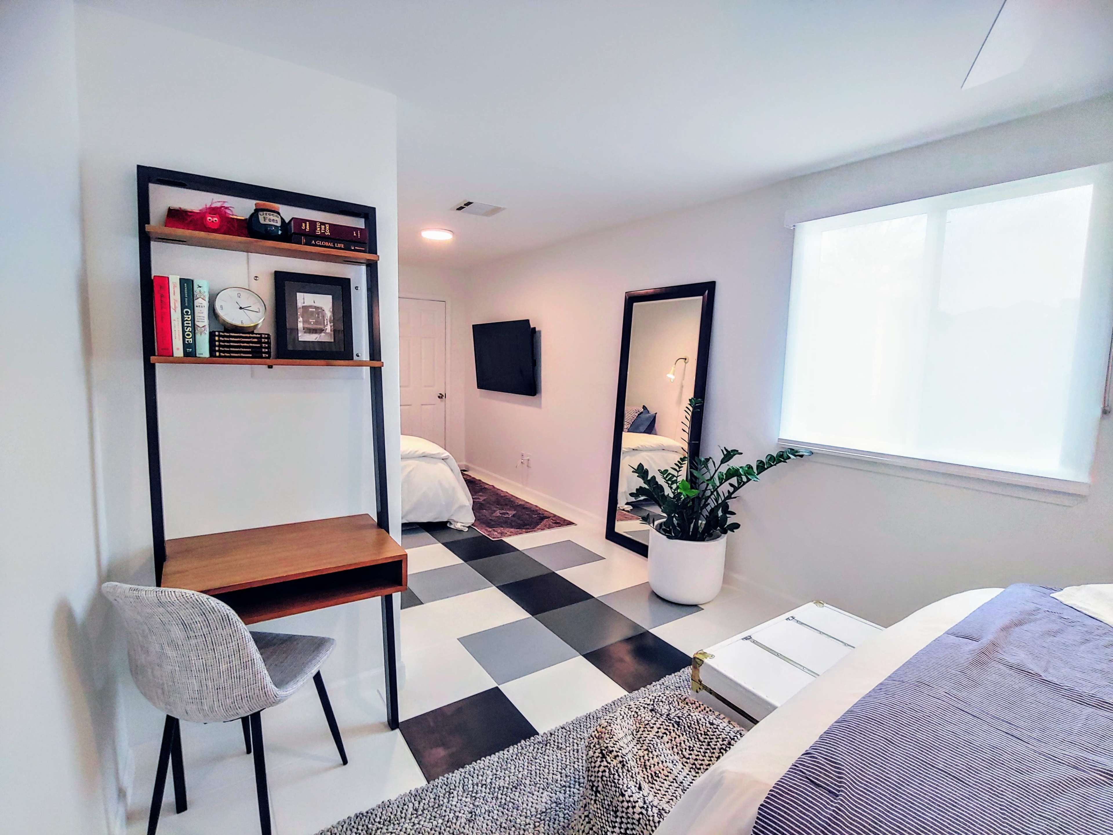 A modern bedroom features a desk with a chair, a mirror, a large plant, and two beds with white linens, set against a black and white checkered floor.