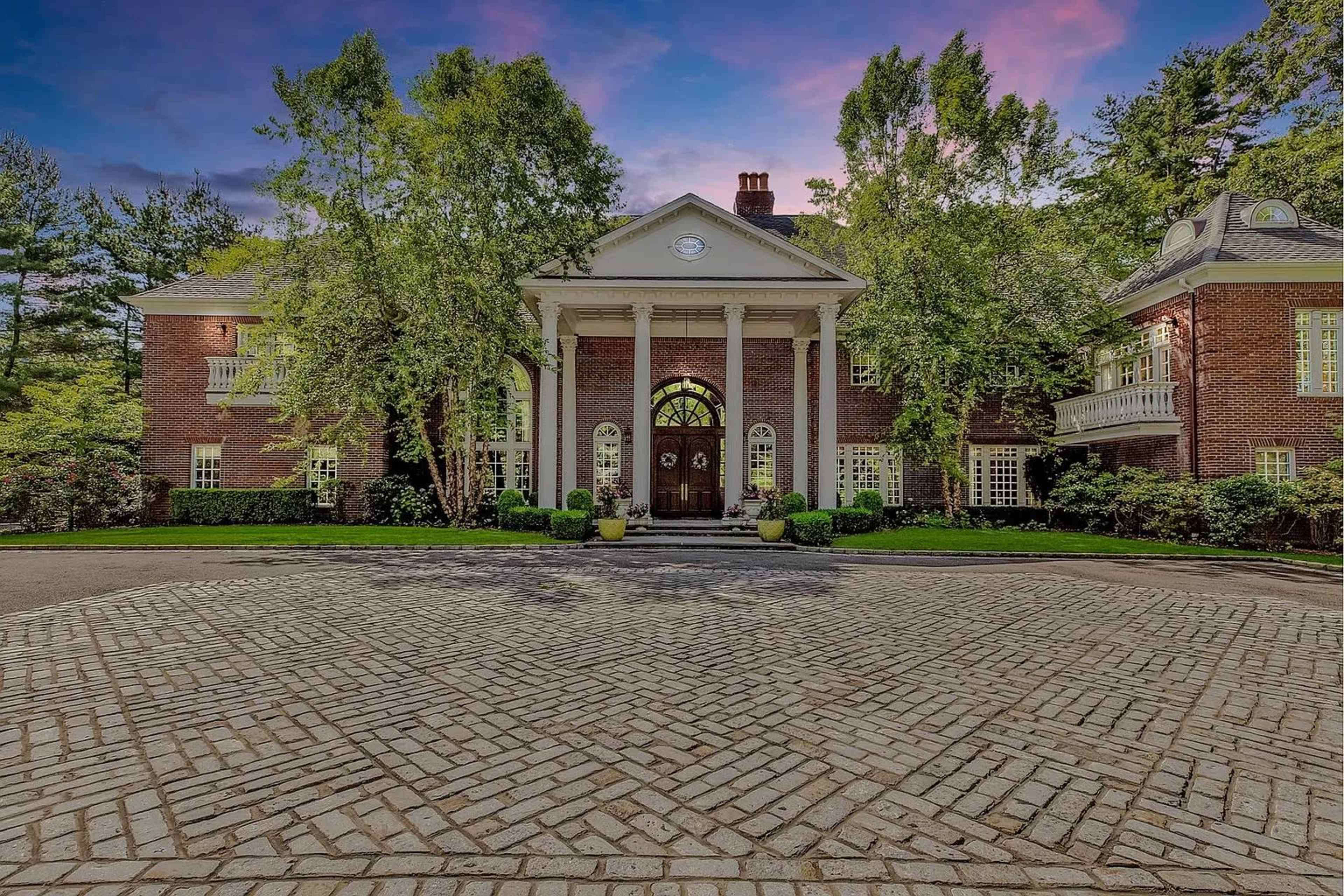A large brick mansion with white columns and a landscaped entrance surrounded by trees.