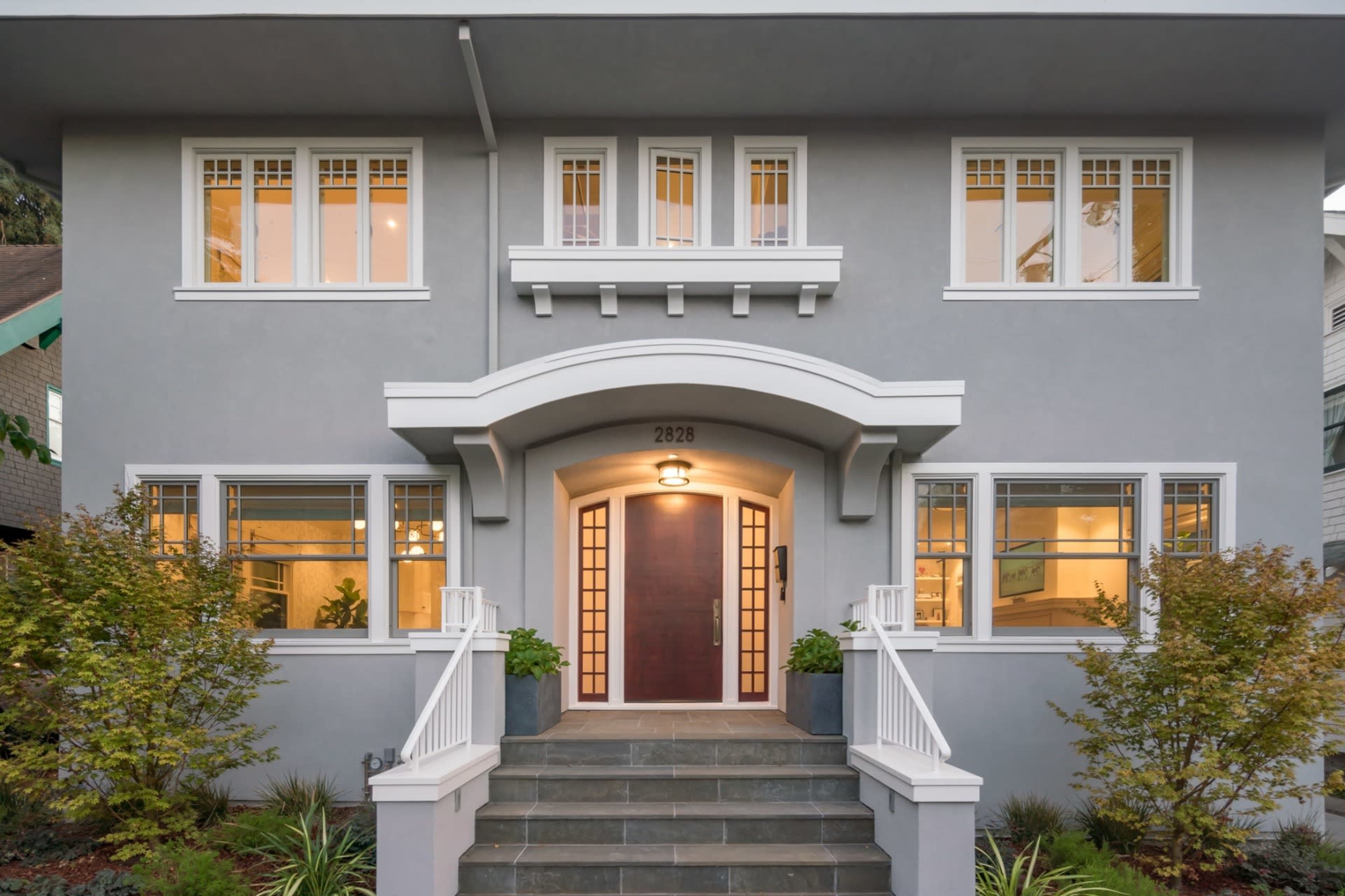 A modern two-story house with a gray exterior, large front windows, and a central wooden door framed by a white archway and stairway.